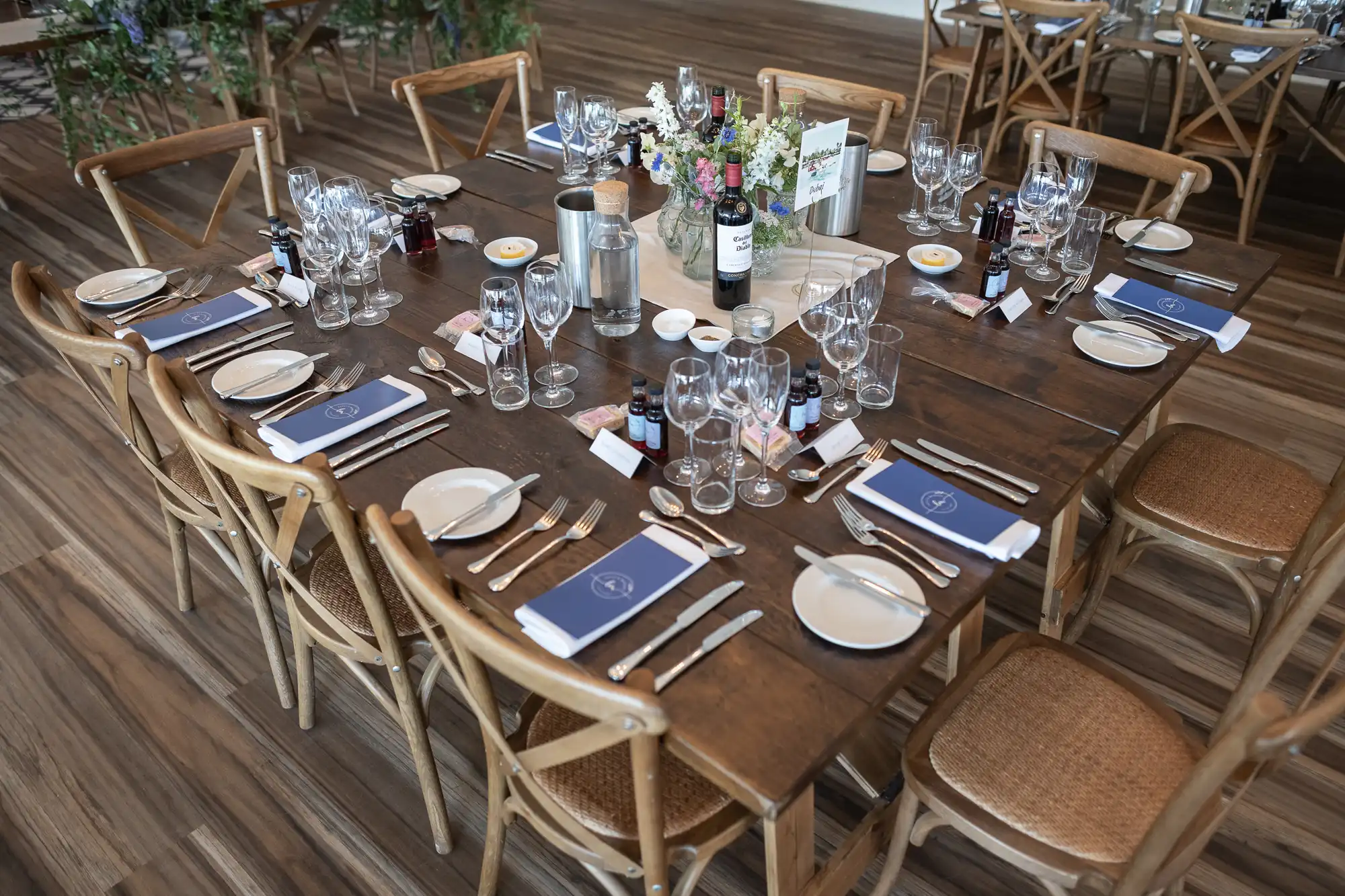 A neatly set dining table with wooden chairs, arranged with glassware, napkins, plates, cutlery, and a floral centerpiece, in a room with wooden flooring.