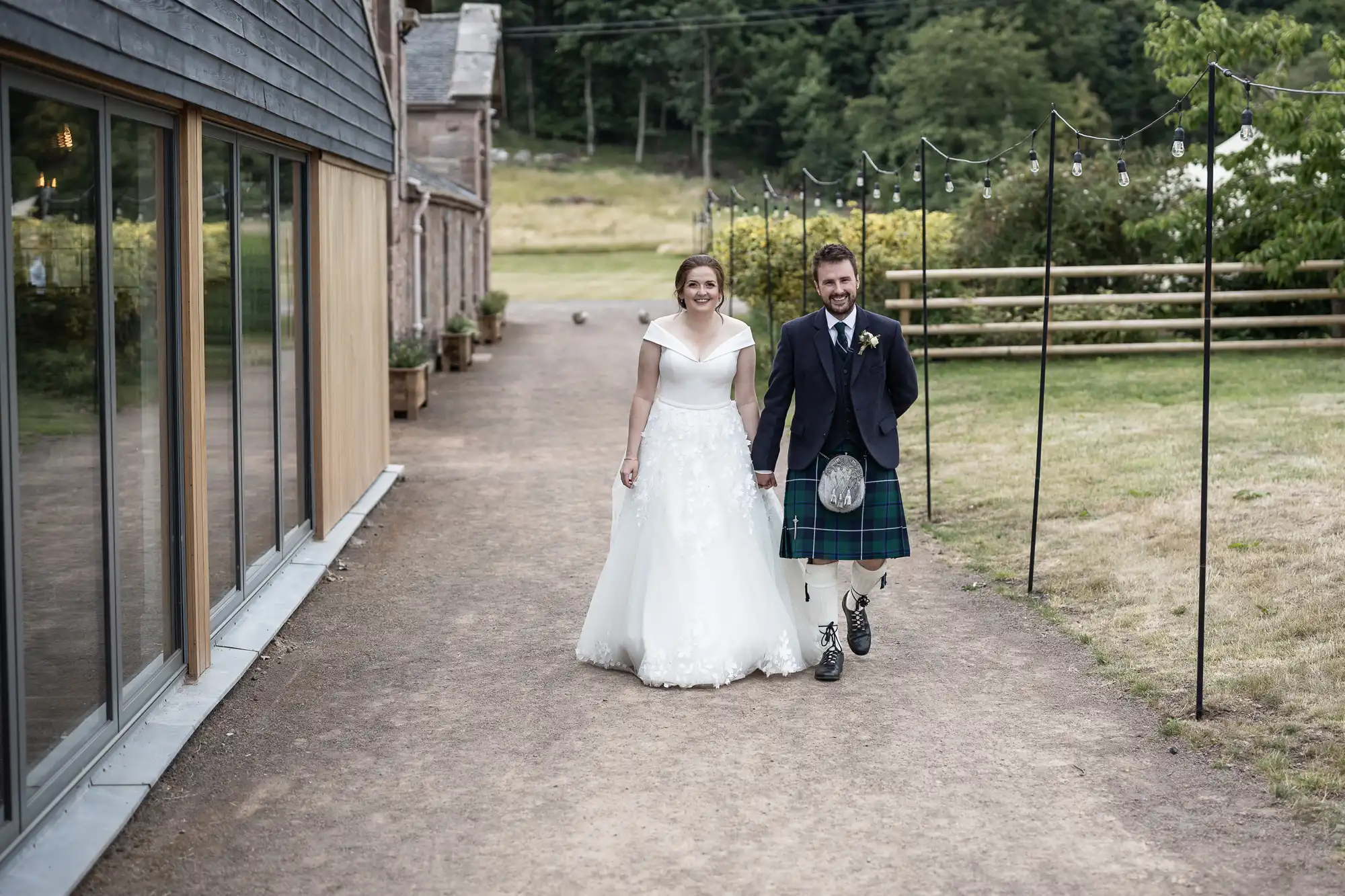 A bride in a white gown and a groom in a kilt walk hand-in-hand on a path beside a building with large windows and outdoor string lights.