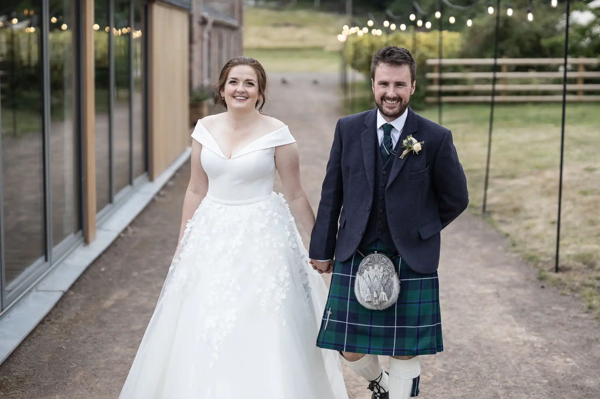 A bride in a white gown and a groom wearing a kilt smile while holding hands, walking outside on a pathway lined with market lights.