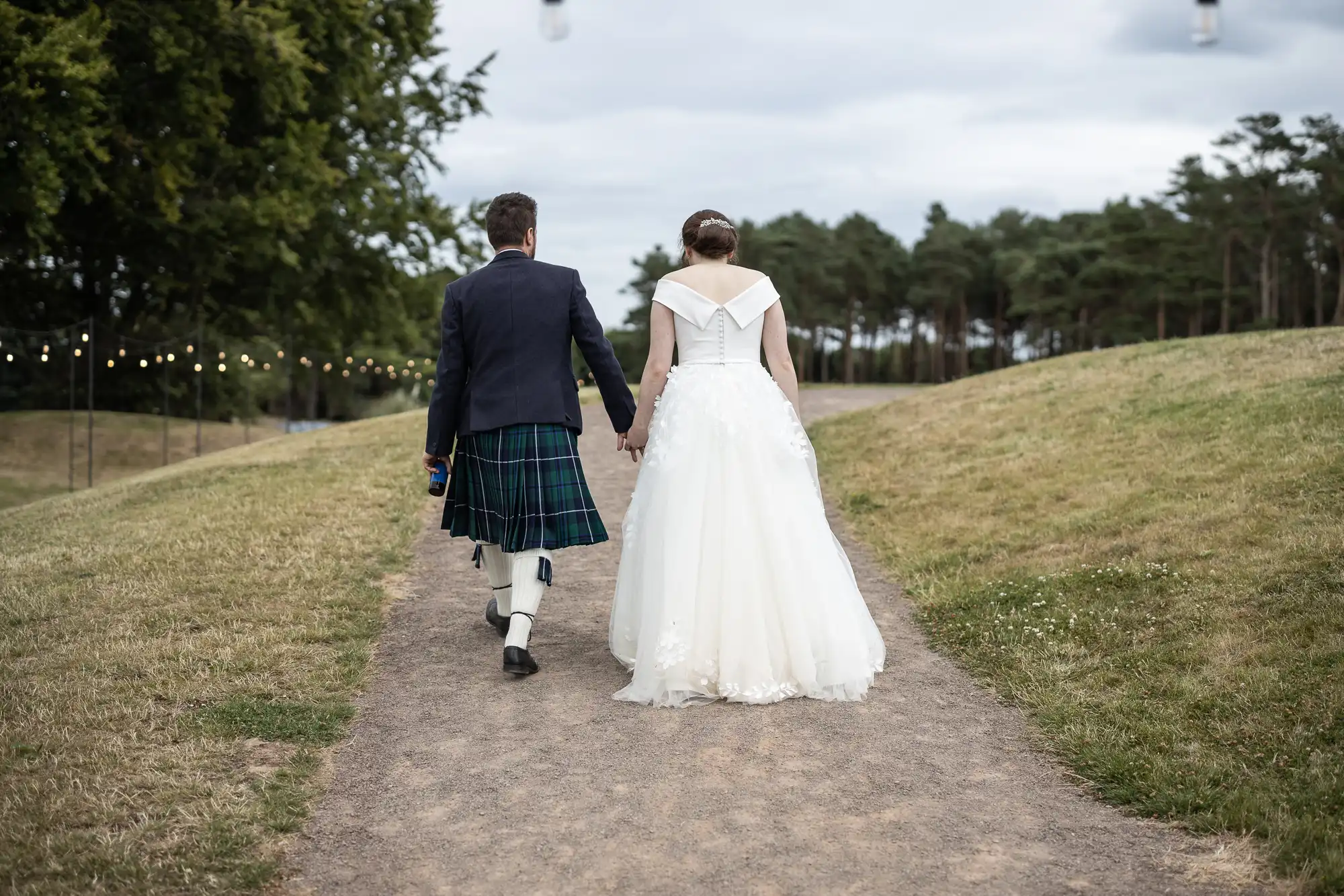 A couple in wedding attire walks hand in hand down a dirt path in a park, with hanging lights and trees in the background.