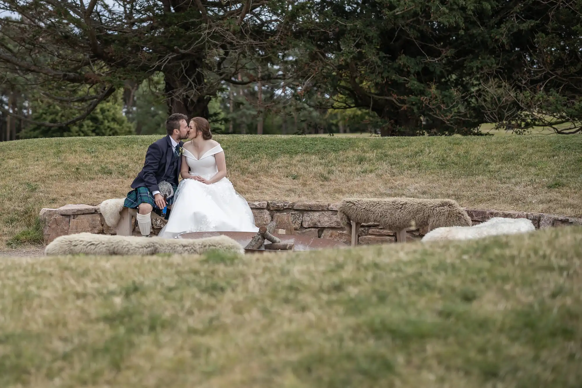 A couple, dressed in wedding attire, sits and kisses on a stone ledge in an outdoor setting with grass and trees in the background.