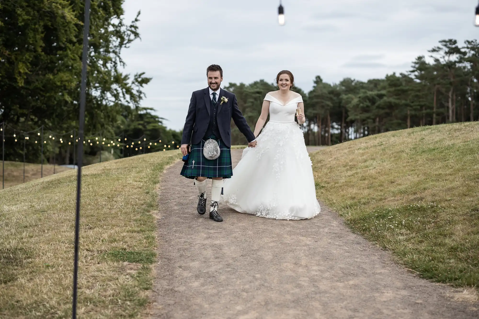 A bride and groom walk hand-in-hand on a path outdoors. The groom wears a traditional Scottish kilt, while the bride wears a white wedding dress. String lights and trees are in the background.