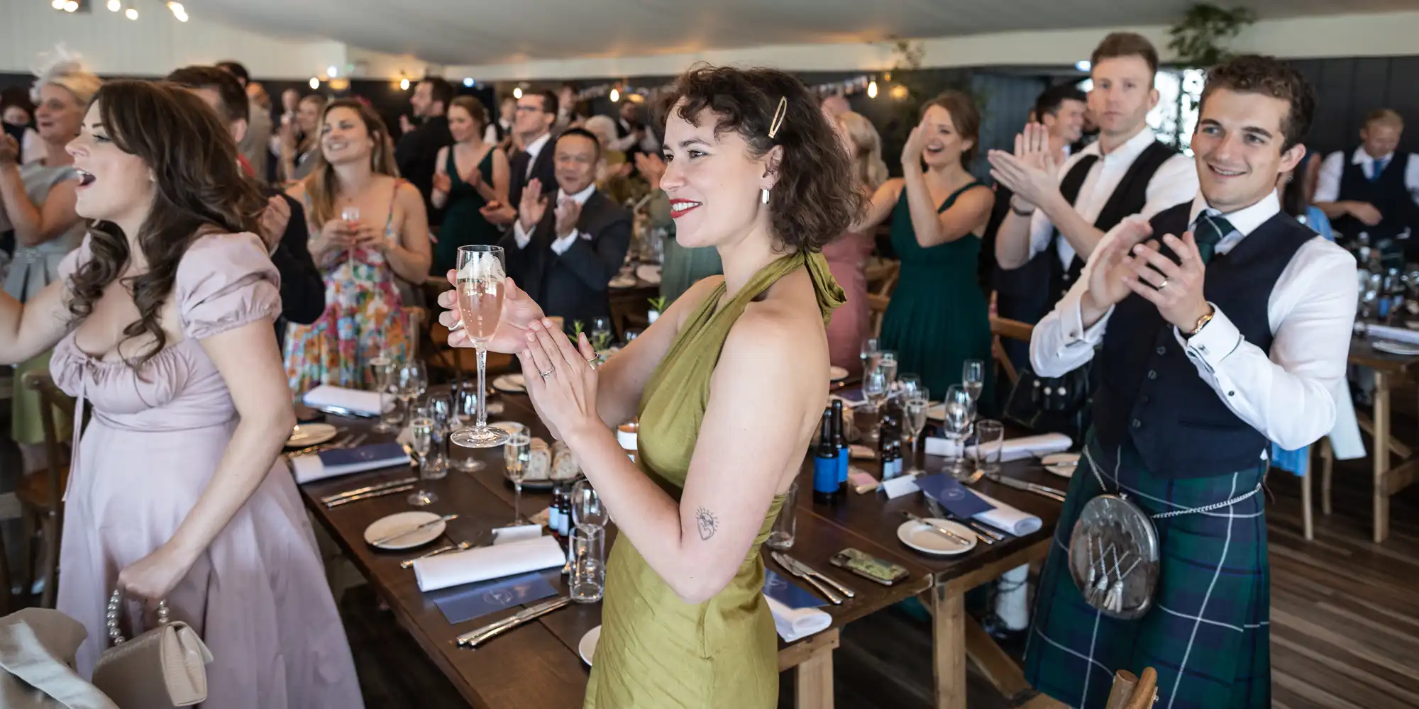 A group of people dressed in formal attire are standing and clapping around dinner tables, with a woman in a green dress holding a glass in the foreground.