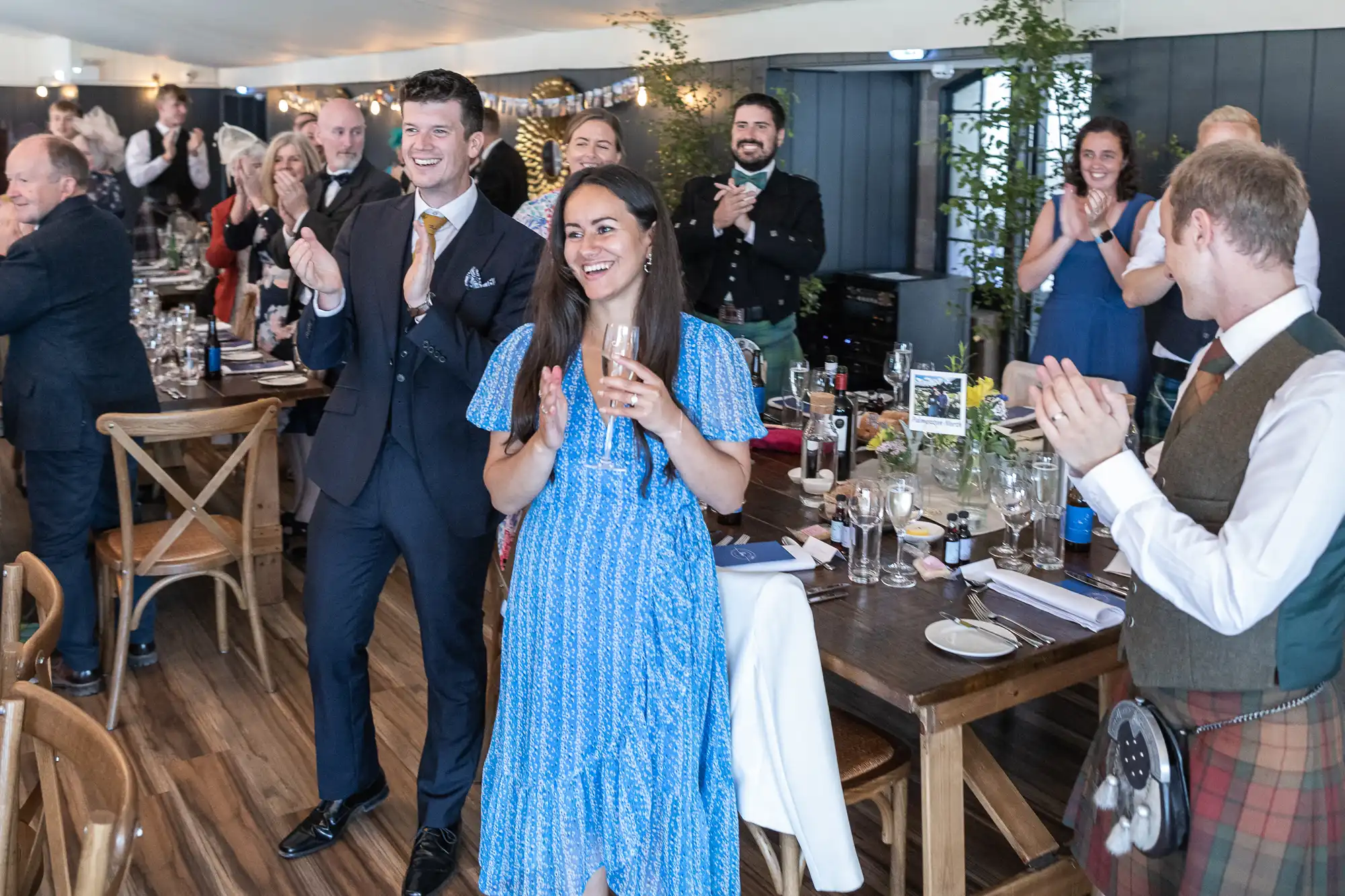 A group of people dressed formally, some in kilts, stand and clap around a dining table in a decorated venue. A woman in a blue dress and a man in a suit are in the foreground.