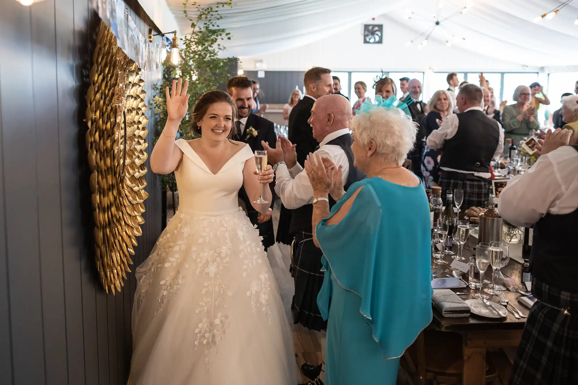 Bride in a white dress holding a glass and waving, standing next to an older man as guests applaud in a warmly lit reception venue.