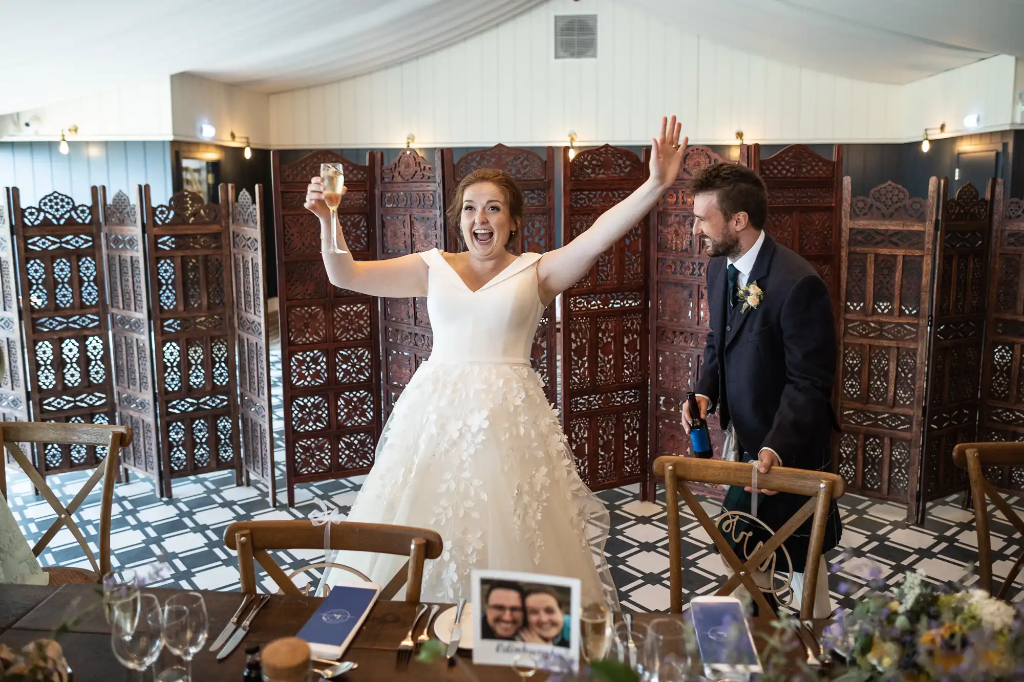 A bride holds up a toast with one arm raised in excitement while a groom stands beside her smiling, in front of an ornately carved wooden screen.