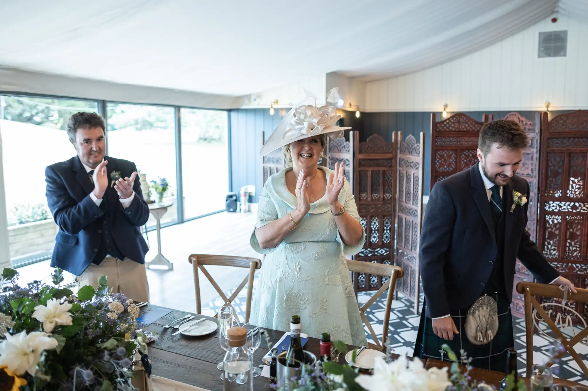 Three people dressed formally are standing by a table, clapping and smiling. They are inside a room with large windows and wooden partitions in the background. The table is decorated with flowers and bottles.