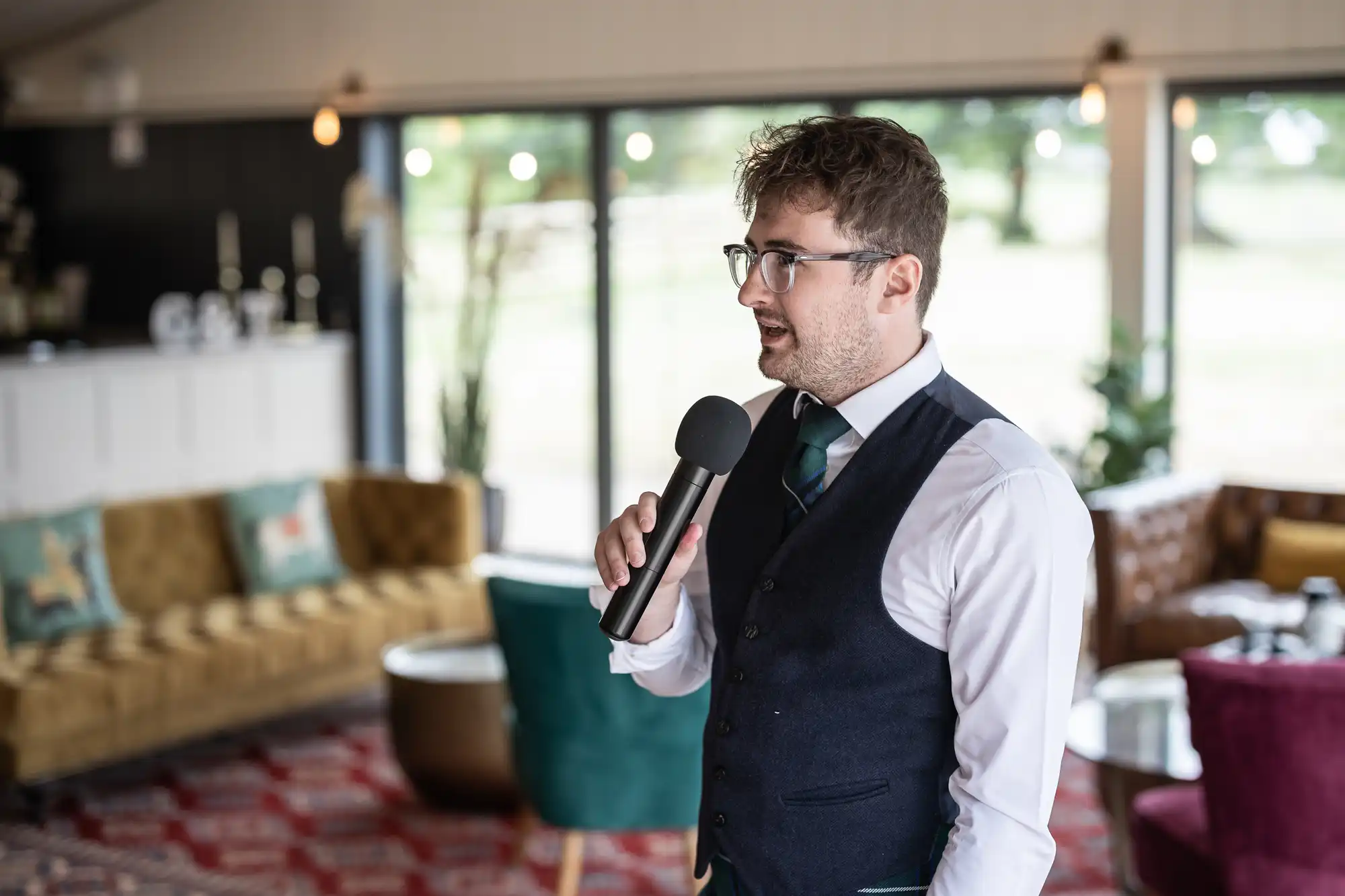 A man in a suit and glasses speaks into a microphone in a modern, well-lit room with large windows, sofas, and decorative cushions.