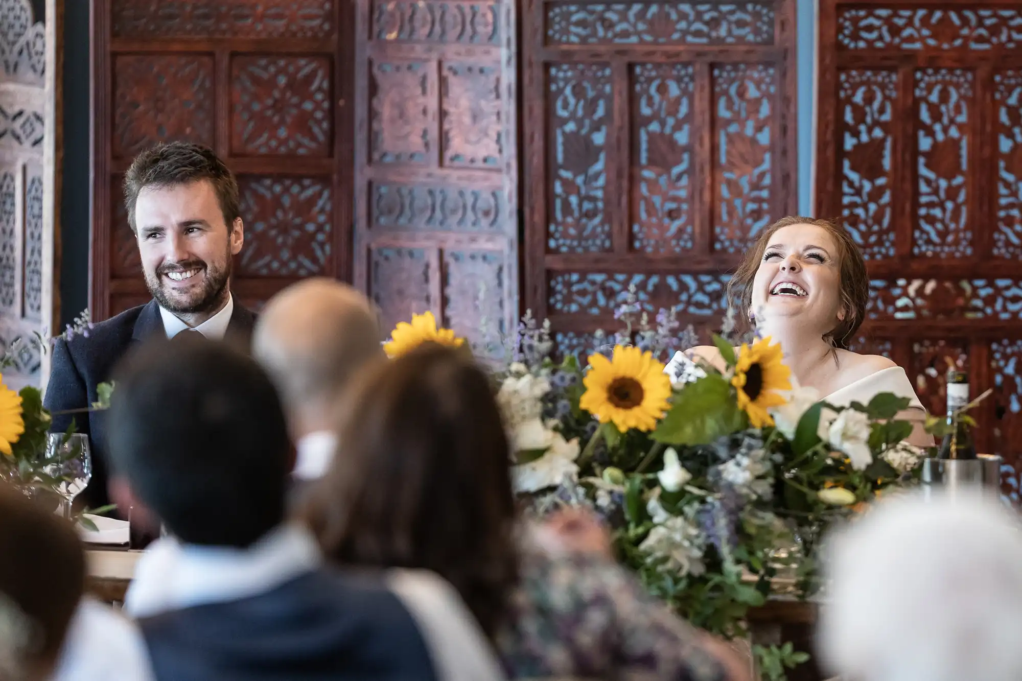 A man and a woman, smiling, are seated at a table adorned with sunflowers and greenery in front of a wooden carved backdrop.