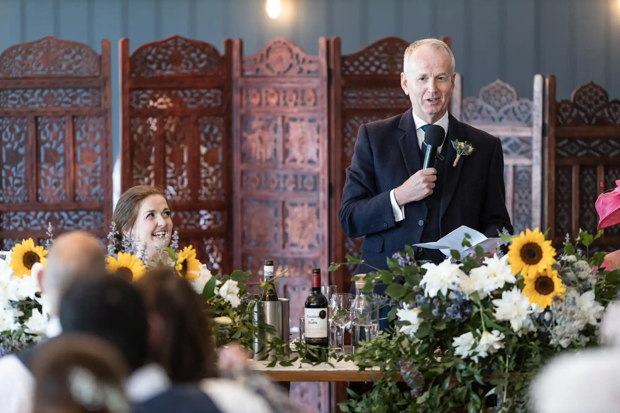 A man in a suit speaks into a microphone at a decorated table with flowers and wine bottles, while a seated woman next to him smiles. An ornate wooden divider is in the background.