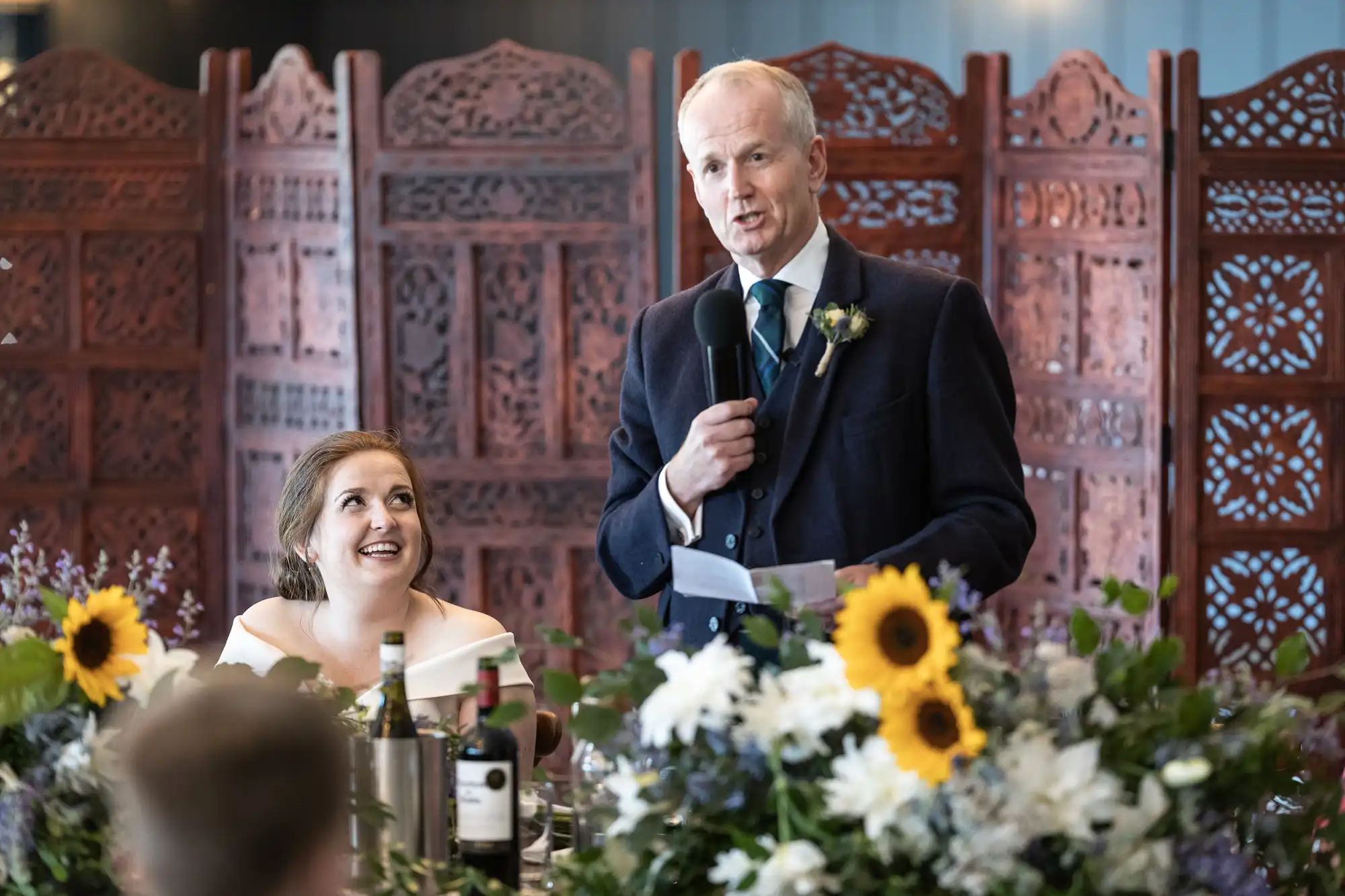 A man in a suit gives a speech using a microphone at a decorated event, while a woman in a white dress sits smiling beside him. Floral arrangements and bottles are visible on the table.