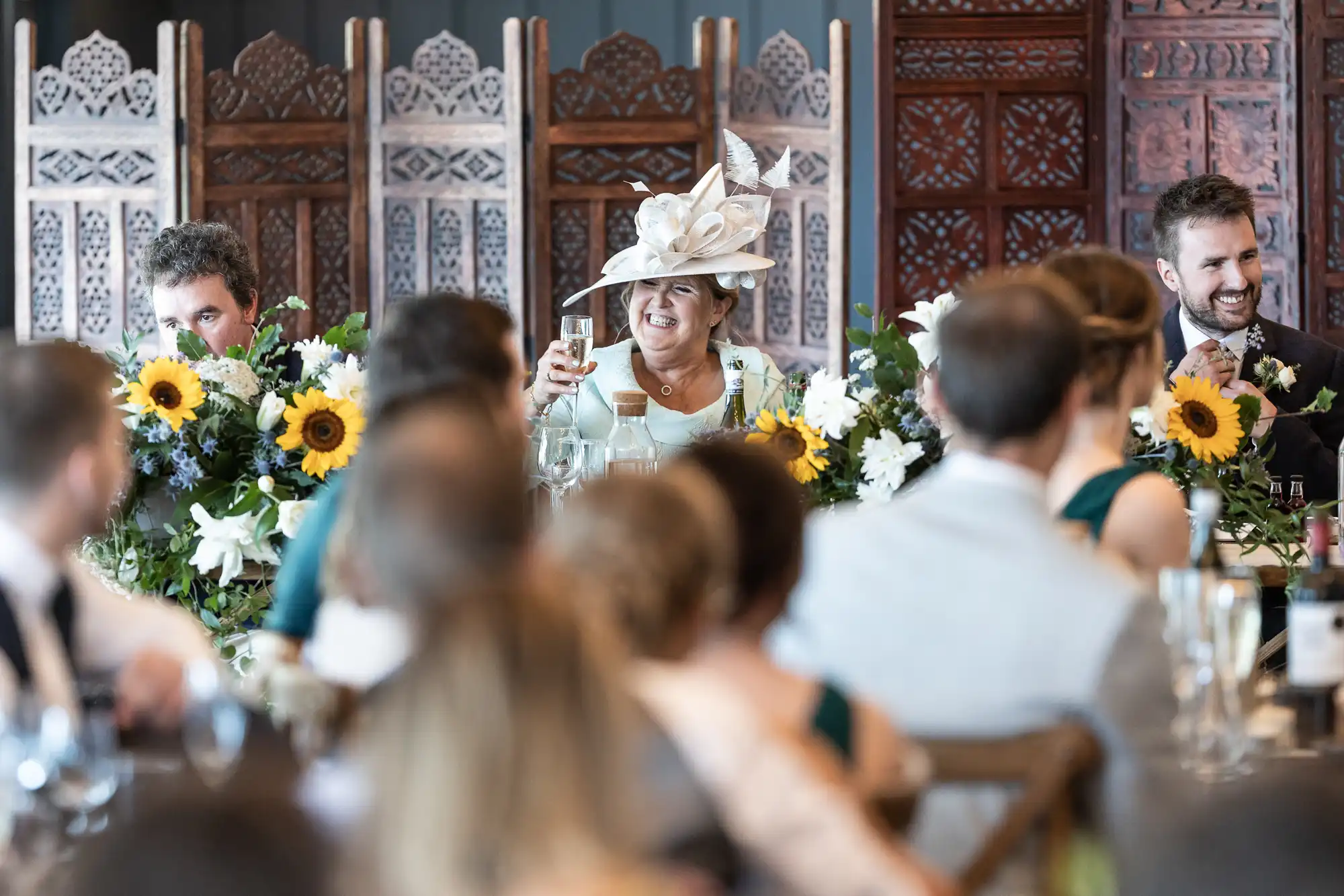 A woman in a white hat raises a glass in a toast at a formal event. She is seated at a table adorned with sunflowers and greenery, surrounded by people dressed in formal attire.