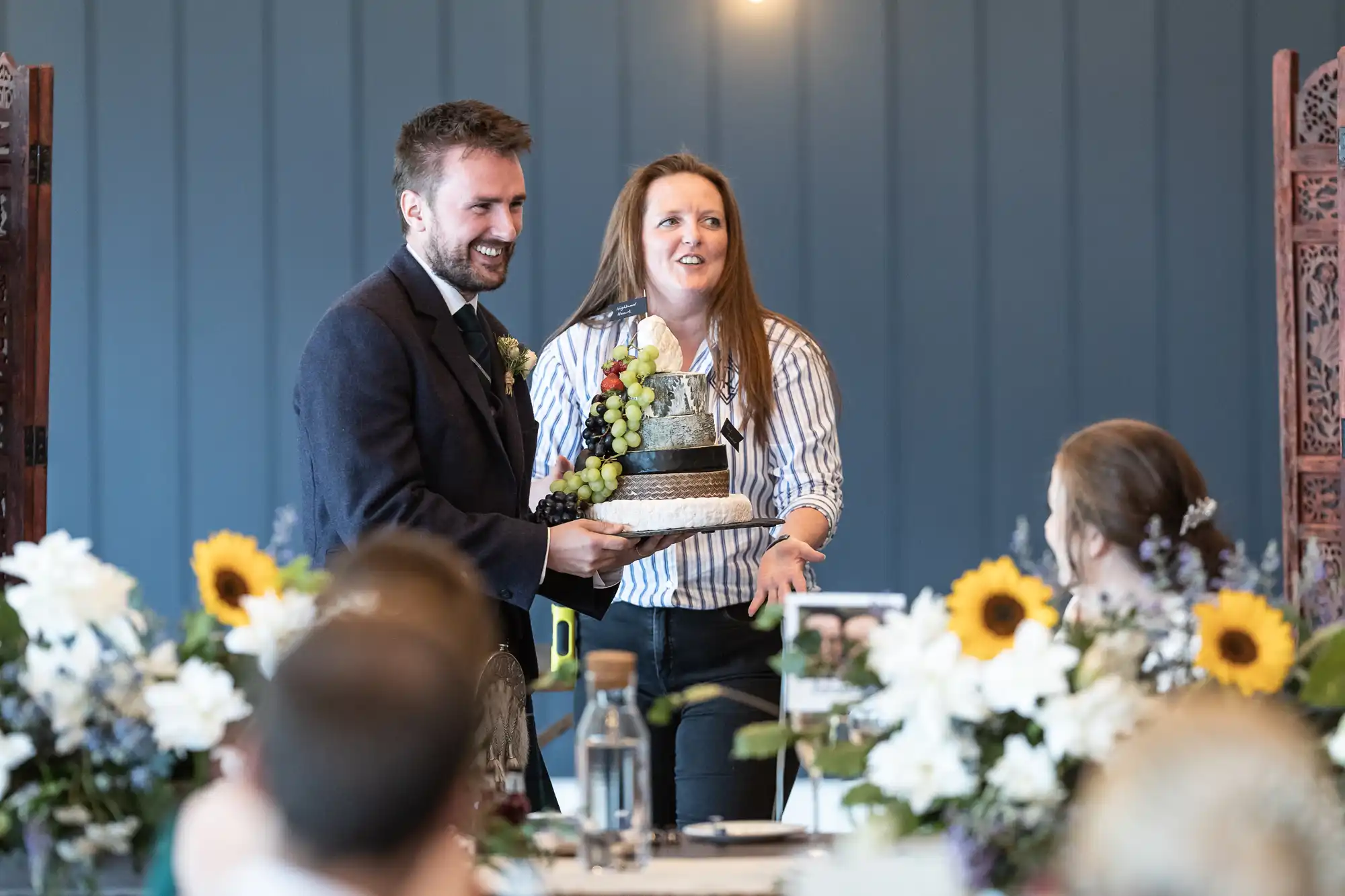 A man and woman stand smiling while holding a decorated cake. They are indoors, surrounded by flowers and people seated at tables.