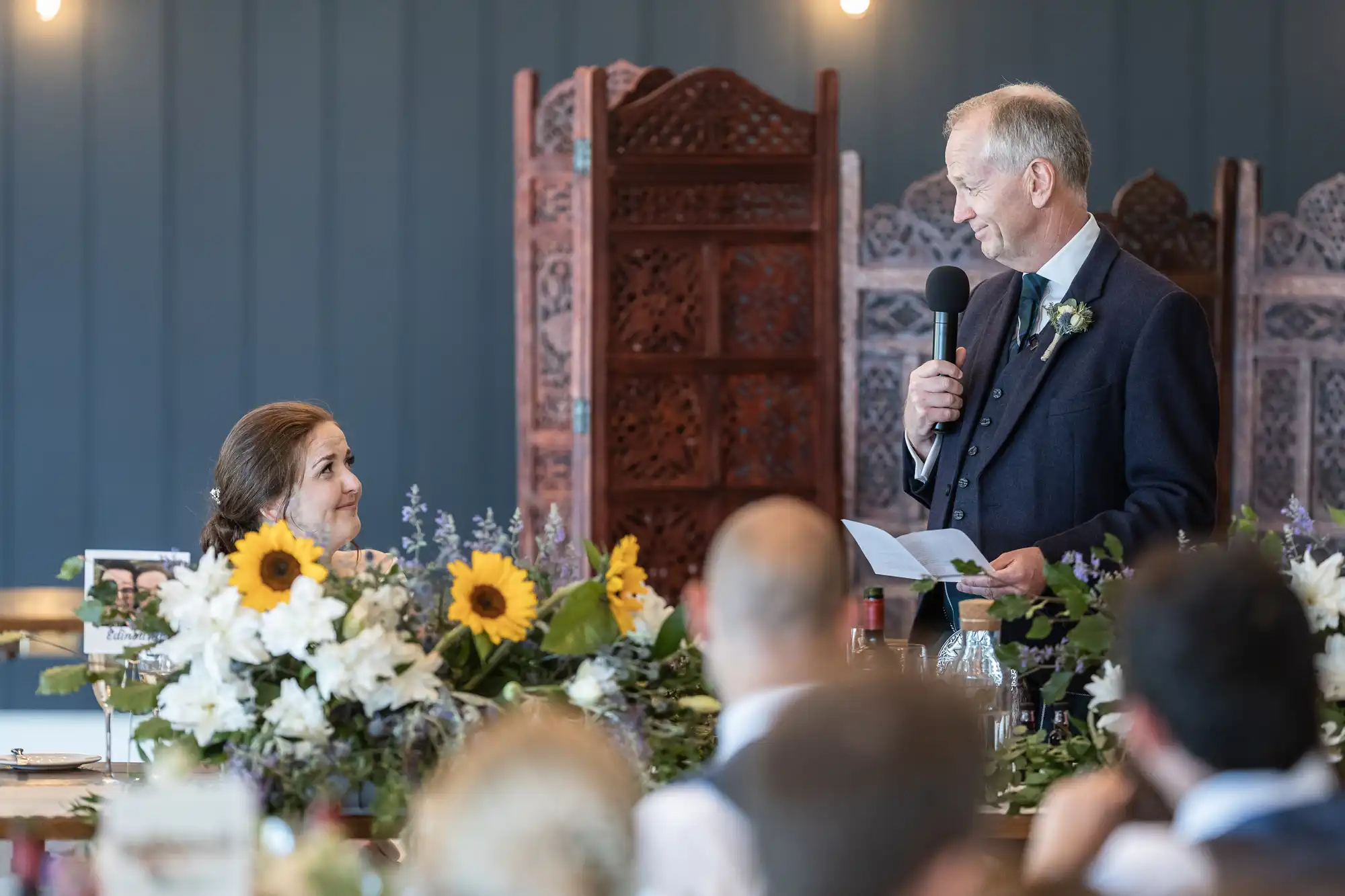 A man with a microphone speaks at a wedding reception as the bride smiles up at him. Floral arrangements with sunflowers decorate the scene.