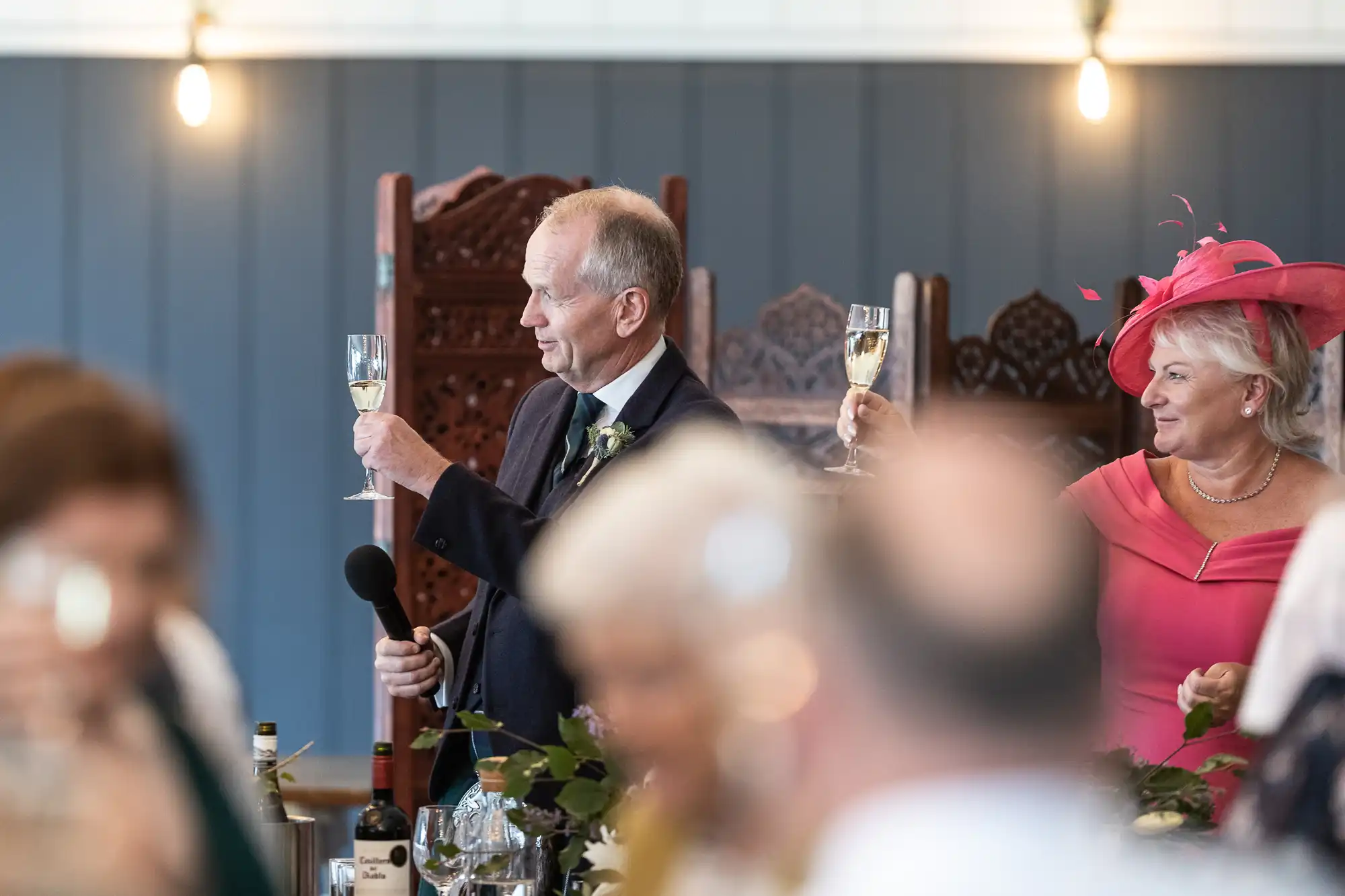 Two people, dressed formally, are raising champagne glasses in a toast at an indoor event. The man, holding a microphone, is beside a decorated table with bottles. The woman is wearing a bright hat.