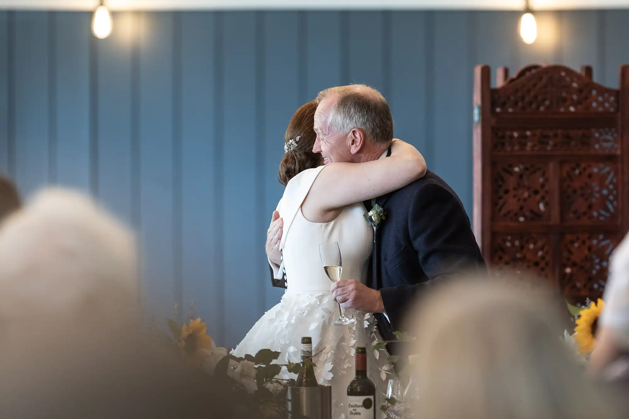 A woman in a white dress and a man in a suit embrace at an indoor event, with a bottle of wine and glasses on the table in the foreground.