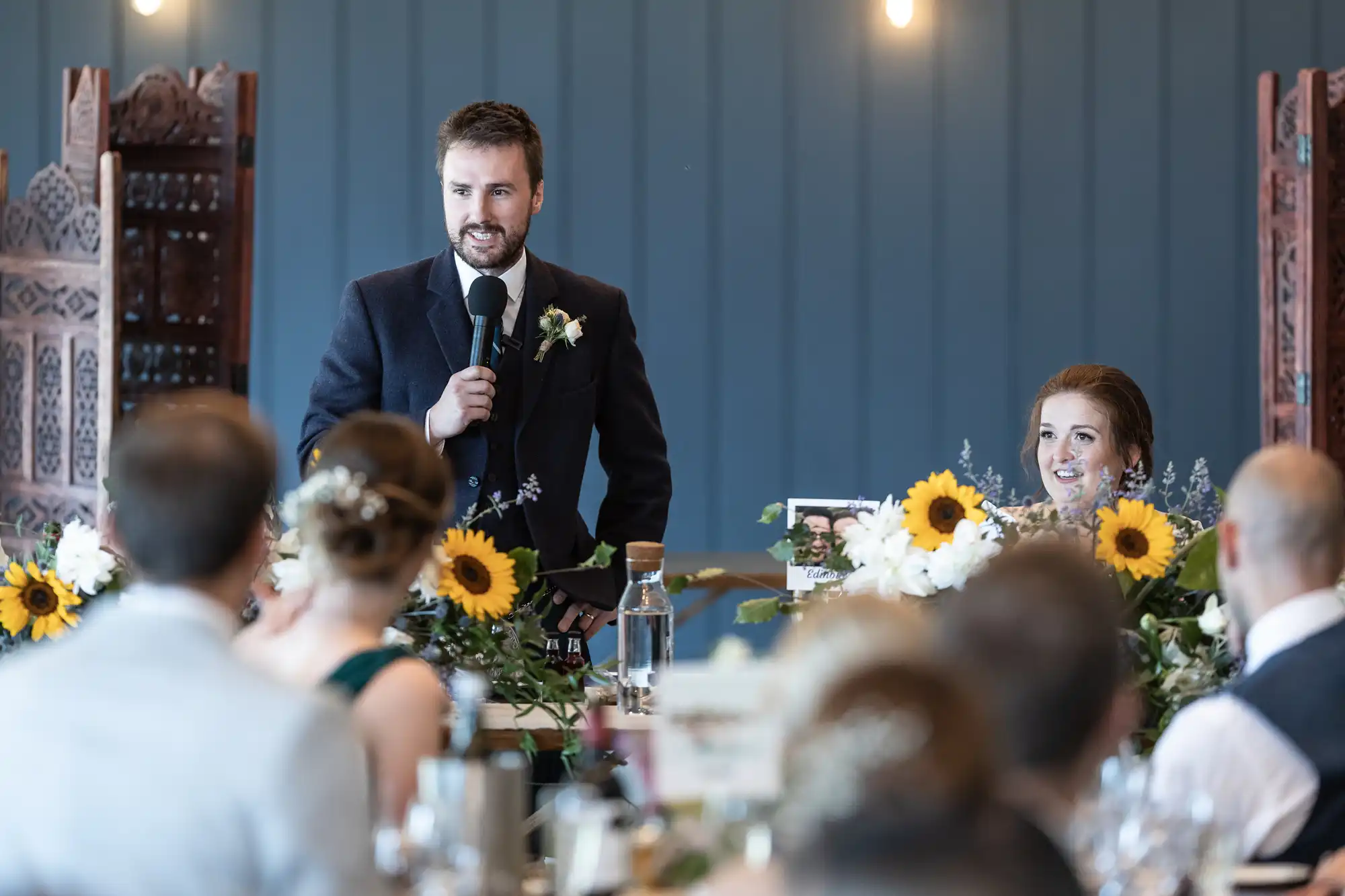 A man in a suit speaks into a microphone at a wedding reception. He stands behind a table adorned with sunflowers and other floral arrangements, with guests seated facing him.