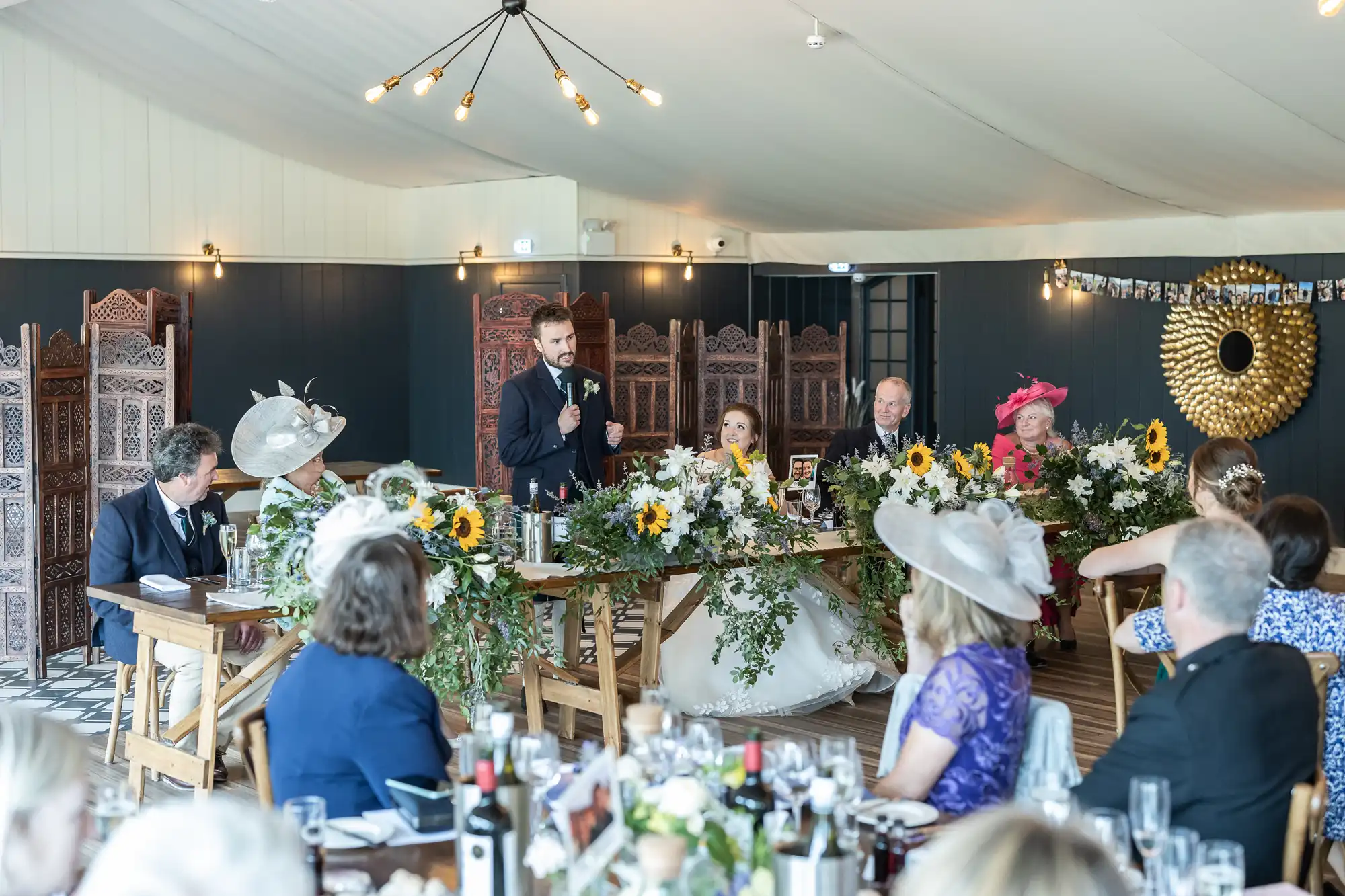 A groom gives a speech at a wedding reception. The bride and several guests sit at a decorated table adorned with sunflowers and greenery. Other guests are seated and watching.