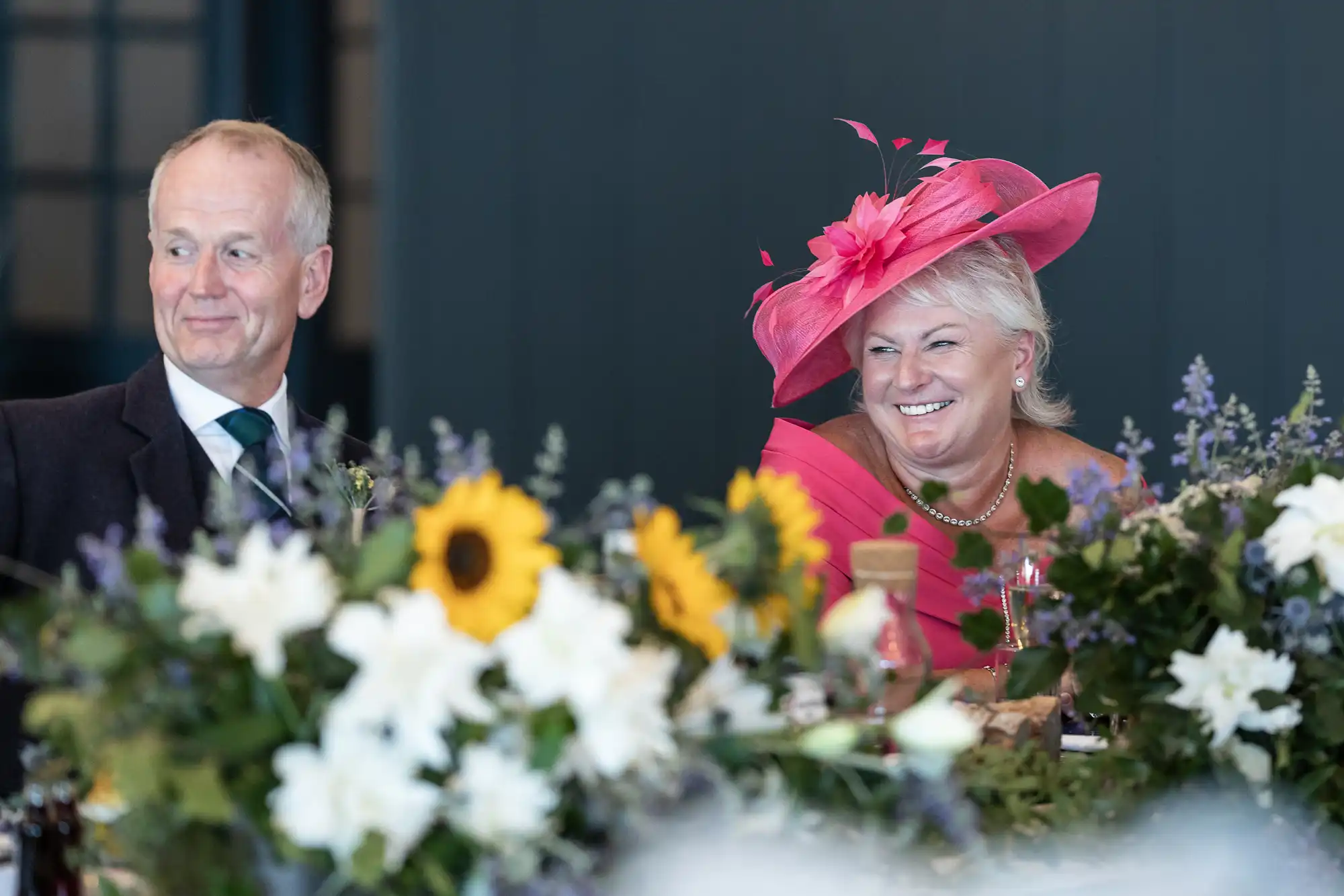 An older man and woman sit at a table adorned with flowers. The woman wears a vibrant pink hat and smiles, while the man looks to the side, both dressed in formal attire.