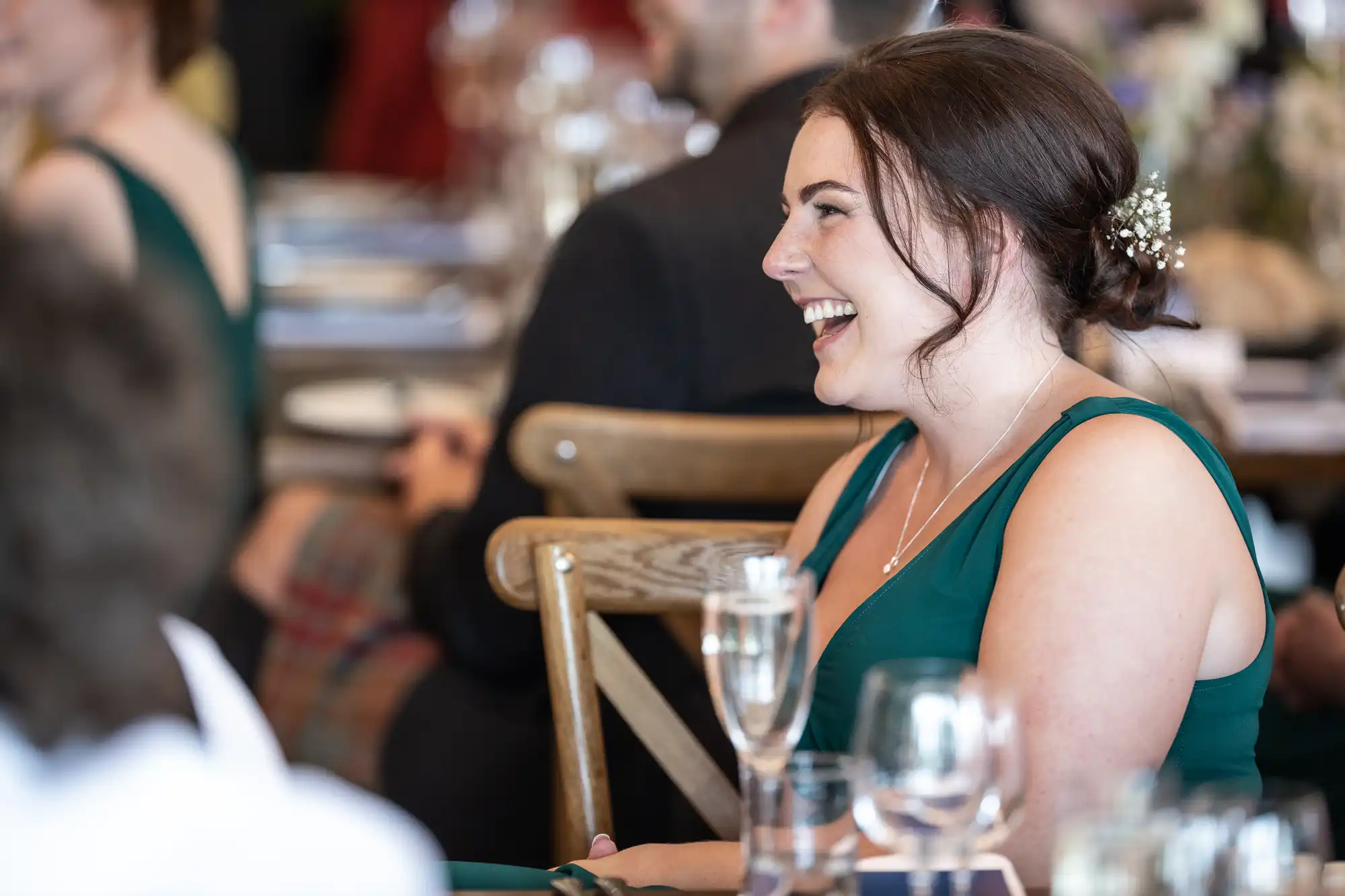 A woman in a green dress smiles while sitting at a table, with a glass of champagne in front of her. Other seated people and blurred background elements are visible.