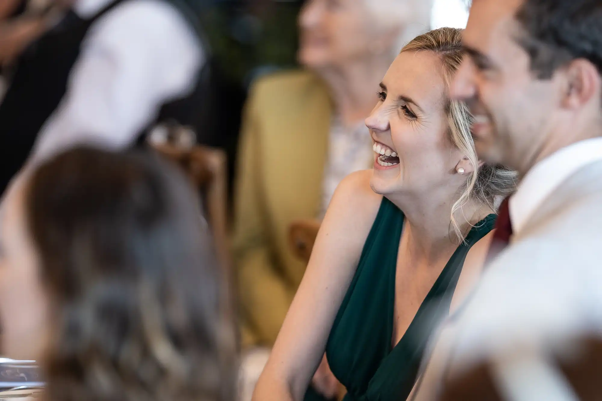 A smiling woman in a green dress and a man in a white shirt with a red tie are seated at an event, with others in the background.