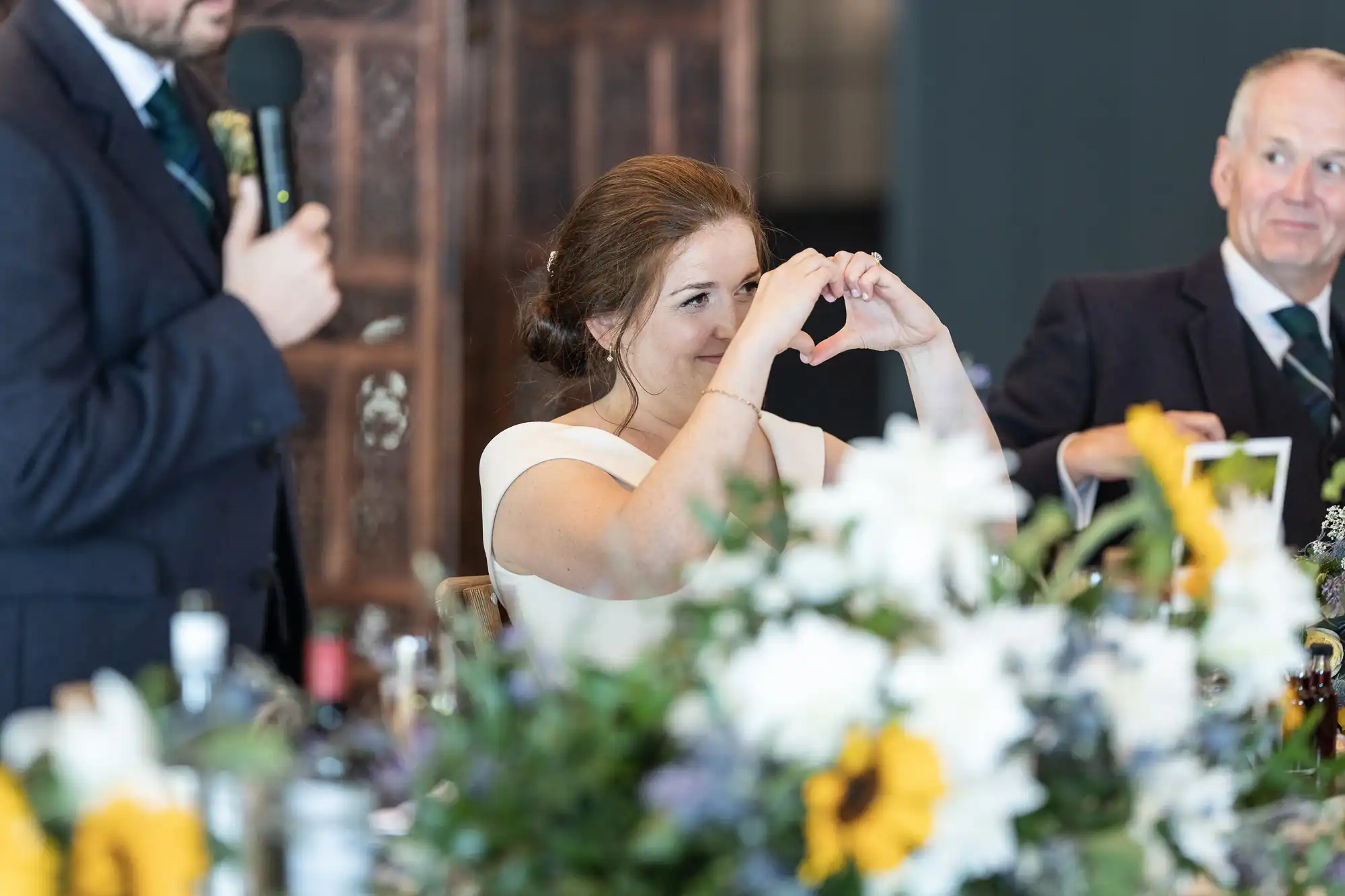 A woman in a white dress makes a heart shape with her hands, sitting at a table with flowers. A man in a suit speaks into a microphone beside her. Another man in a suit is seated on her other side.