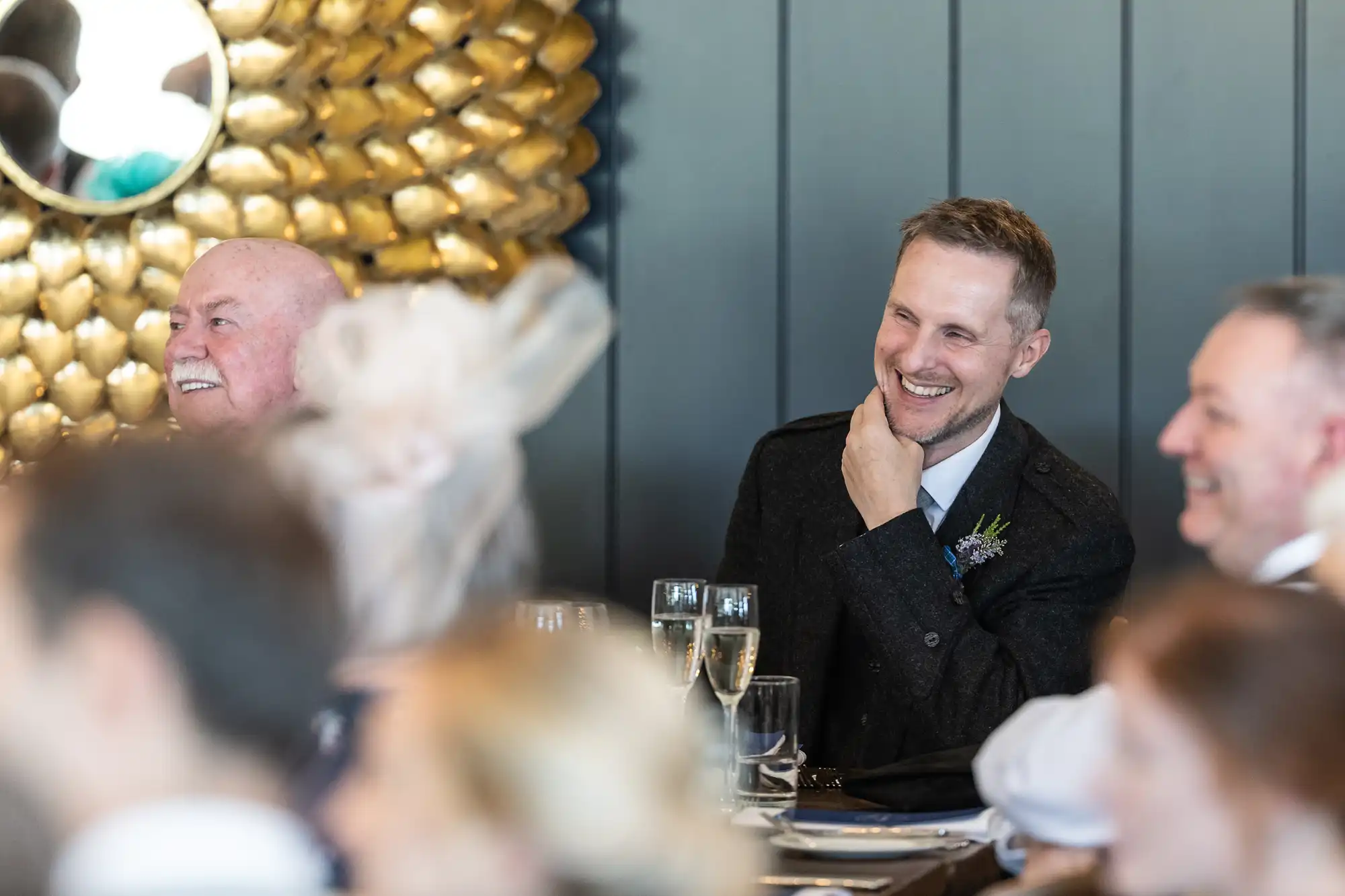 Two men in formal attire smile and laugh at an indoor event, with blurred attendees in the foreground and a decorative golden wall in the background.