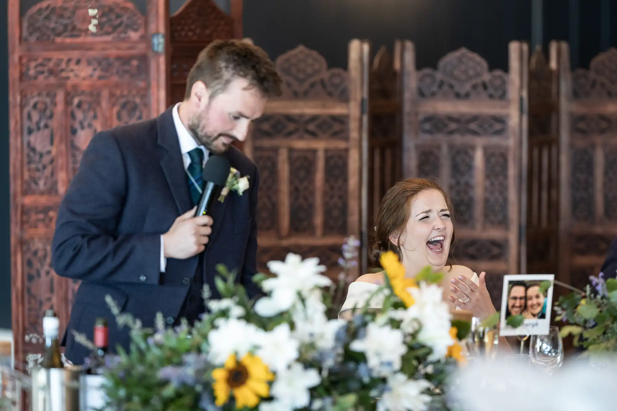 A man in a suit holds a microphone while a woman laughs heartily at a table with flowers and drinks. The background features ornate wooden panels.