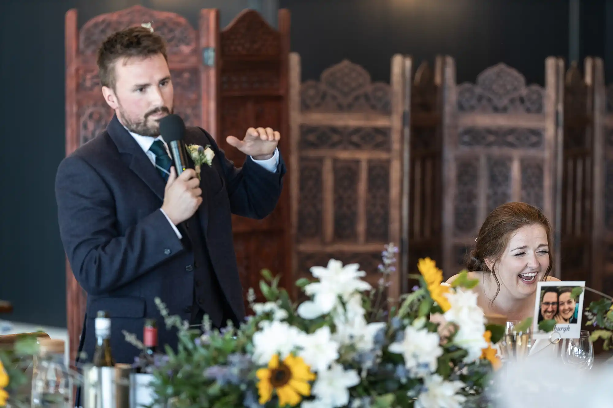 A man in a suit speaks into a microphone at an event. A woman beside him is sitting and laughing while holding a picture. Flowers and wine bottles are on the table in front of them.