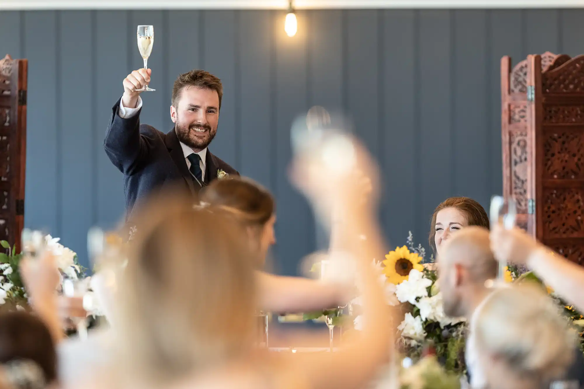 A man in a suit raises a glass in a toast at a wedding reception while guests raise their glasses in response. Flowers and warm lighting are present in the background.