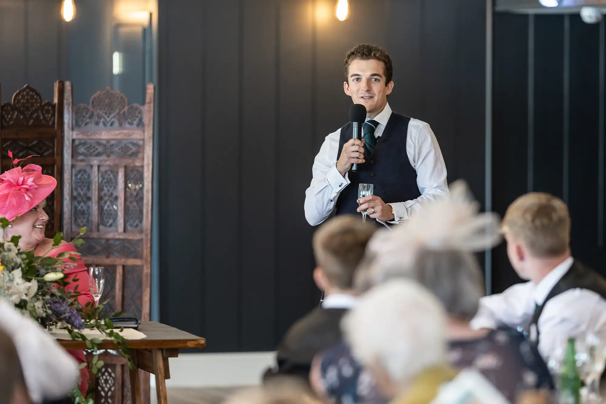 A man in formal attire is speaking into a microphone, holding a glass, while standing in front of a seated audience.