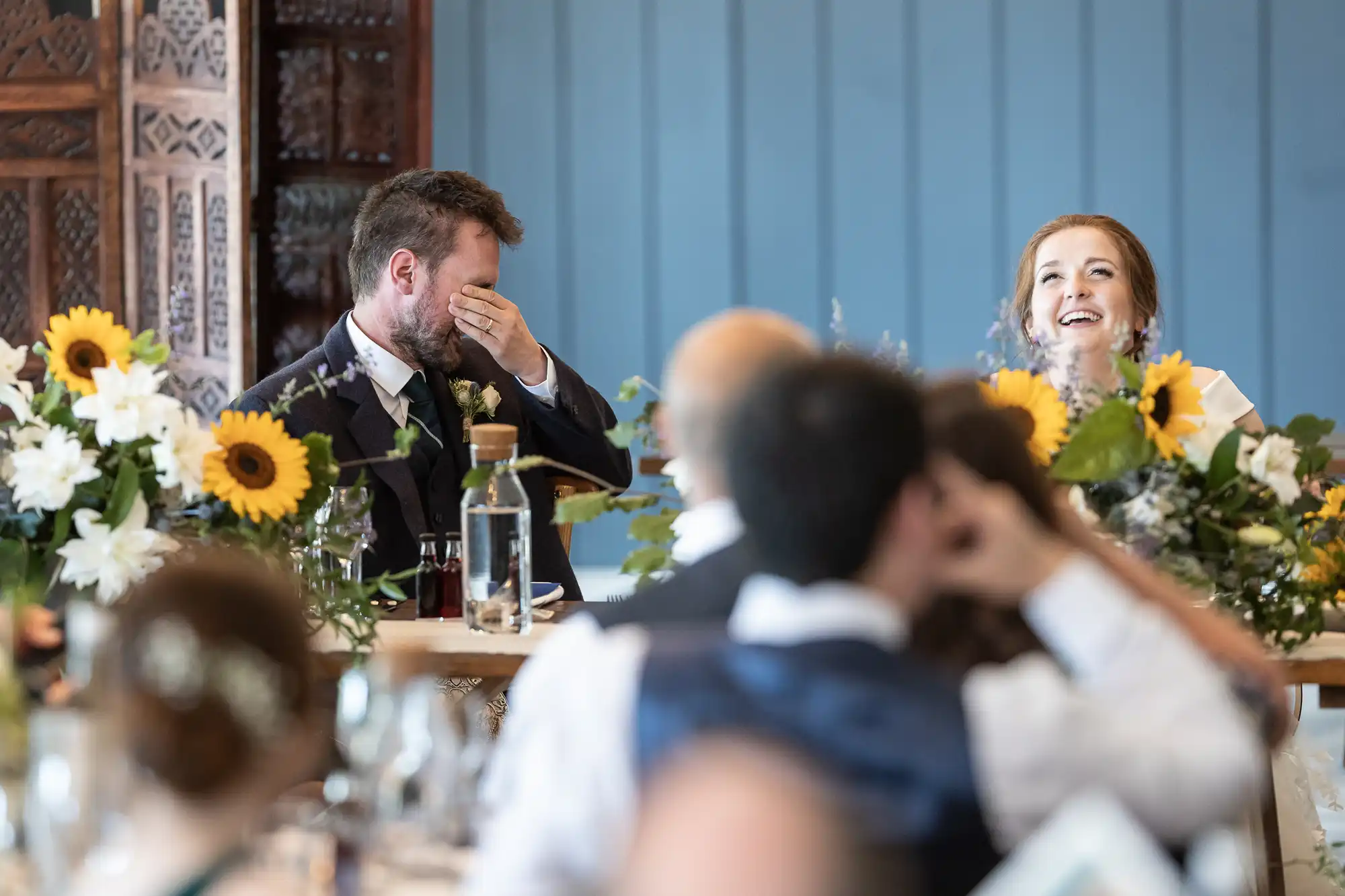 A bride and groom sit at a decorated table with sunflowers and laugh during a wedding reception. The groom covers his face with his hand while the bride looks up and smiles. Guests are seated around them.