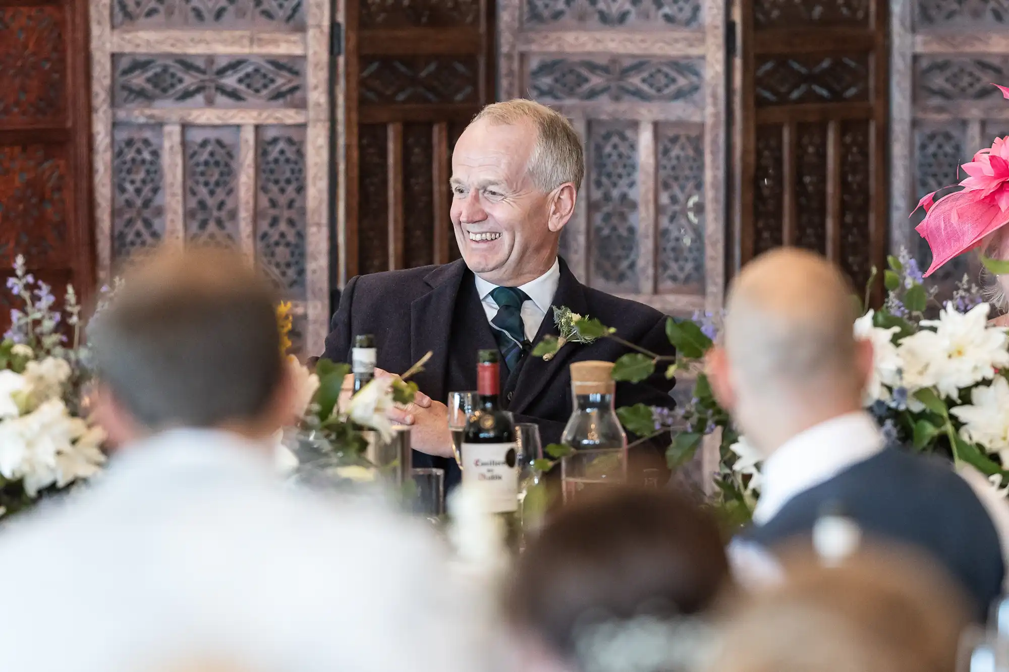 A man in a suit sits at a table, smiling and surrounded by floral arrangements. Other people are seen facing him in the foreground.