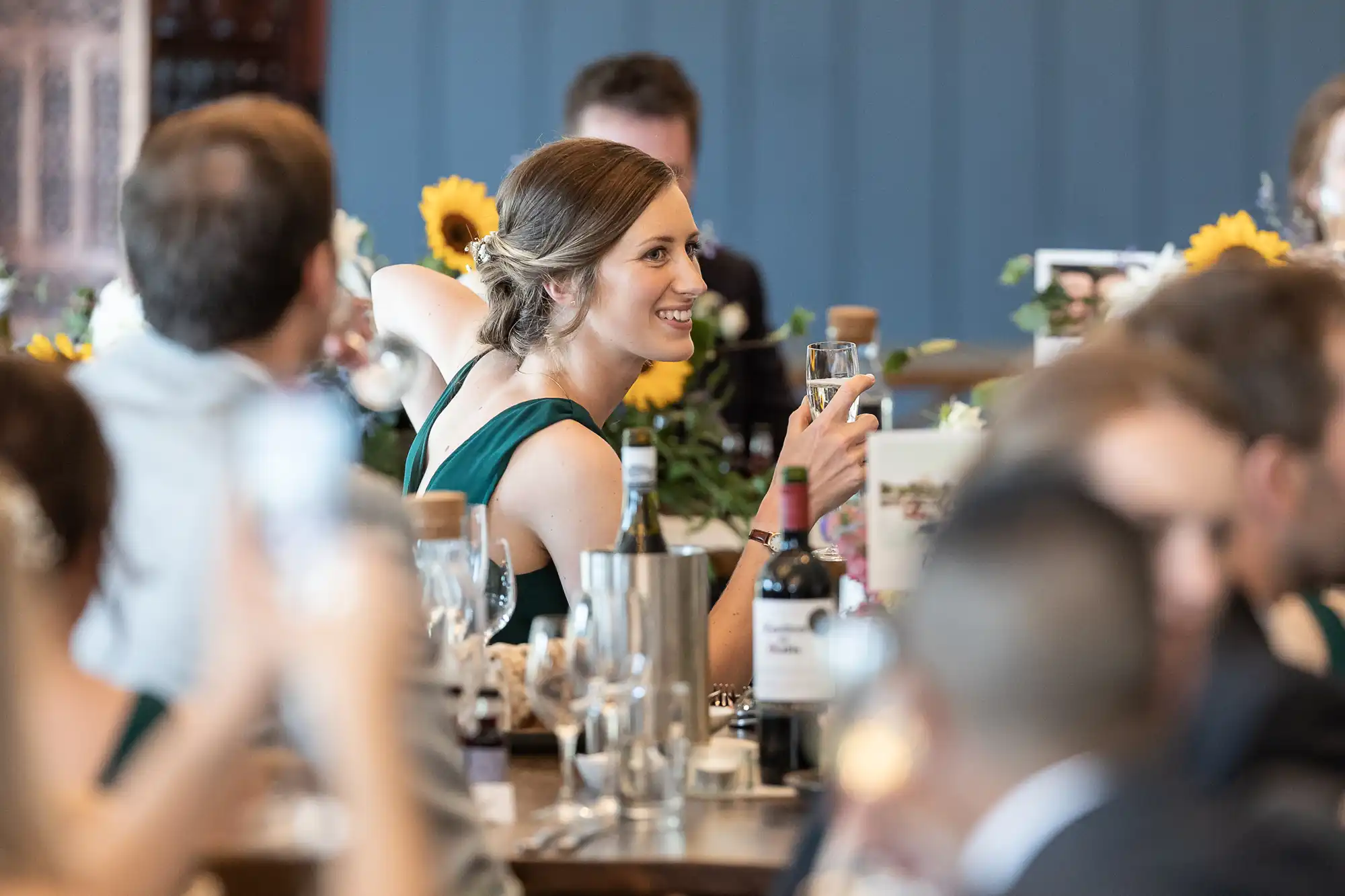 A smiling woman holds a glass while seated among others at a dining table adorned with bottles, glasses, and sunflower decorations.