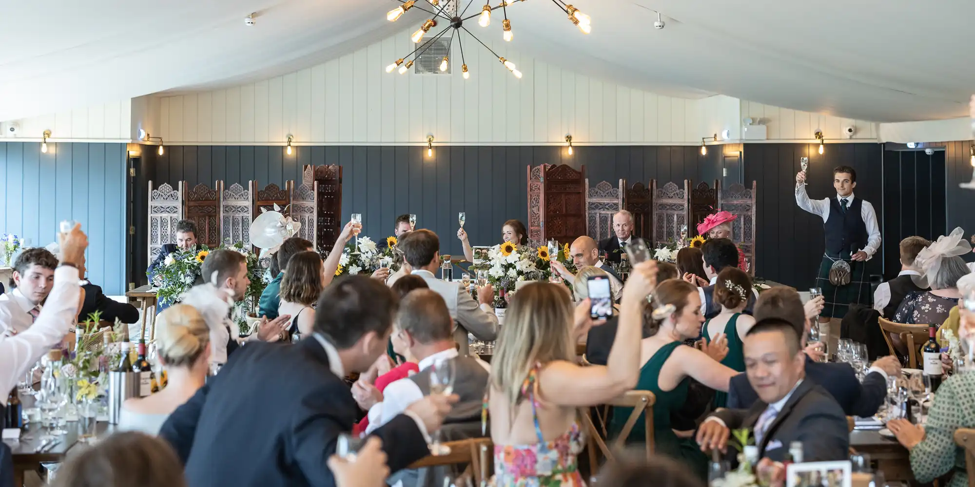 A wedding reception with guests raising glasses in a toast. The room is decorated with flowers and various lights, and a man standing near the head table appears to be making a speech.