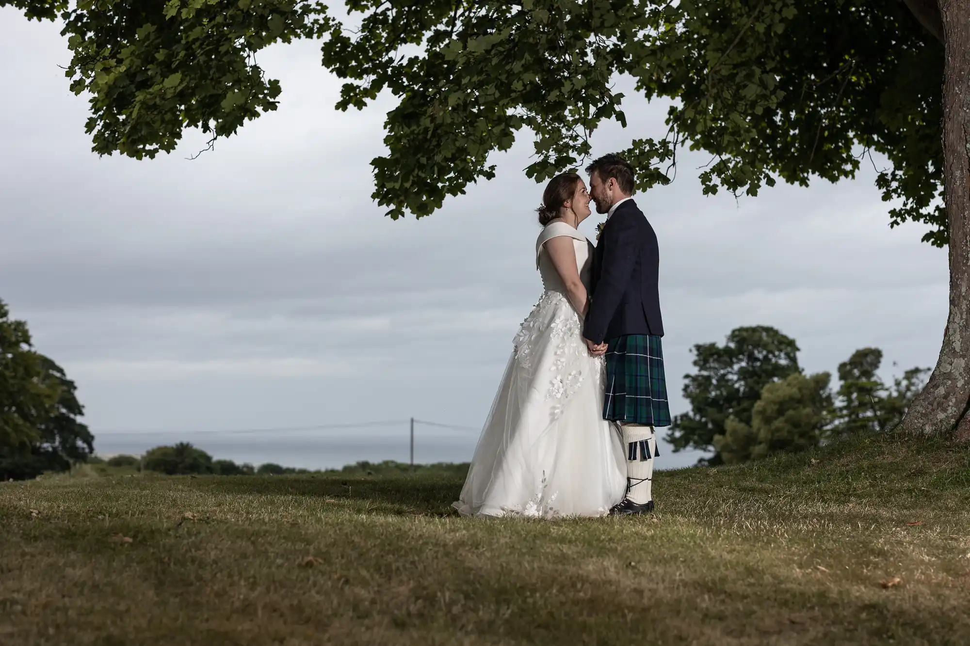 A bride and groom face each other holding hands under a large tree on a grassy lawn, with a cloudy sky and distant view of water in the background. The groom wears a kilt and the bride a white gown.