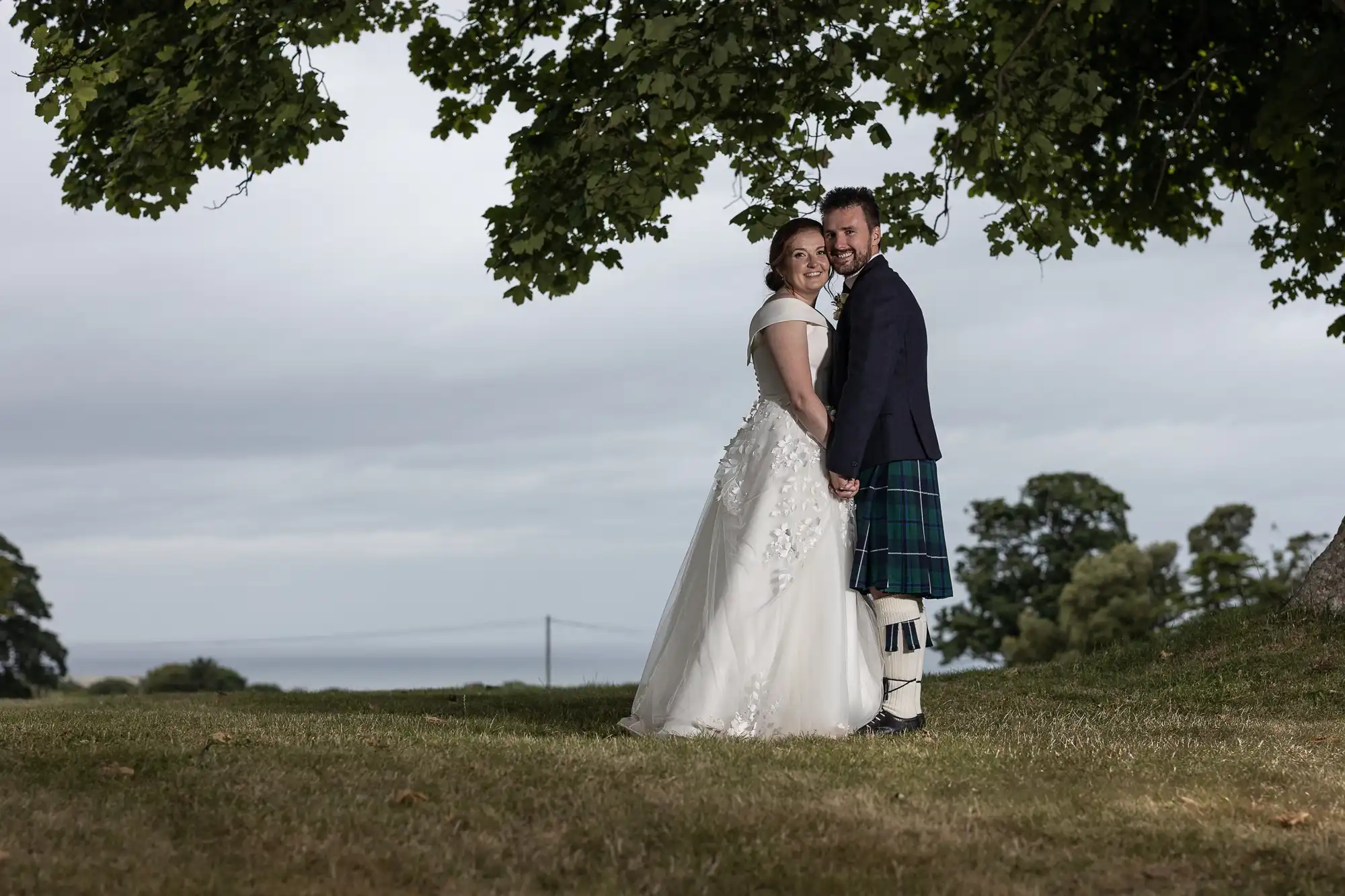 A bride and groom stand outdoors on a grassy area under a tree, holding hands and smiling. The groom wears a kilt, and the bride is in a white gown. A cloudy sky and body of water are in the background.