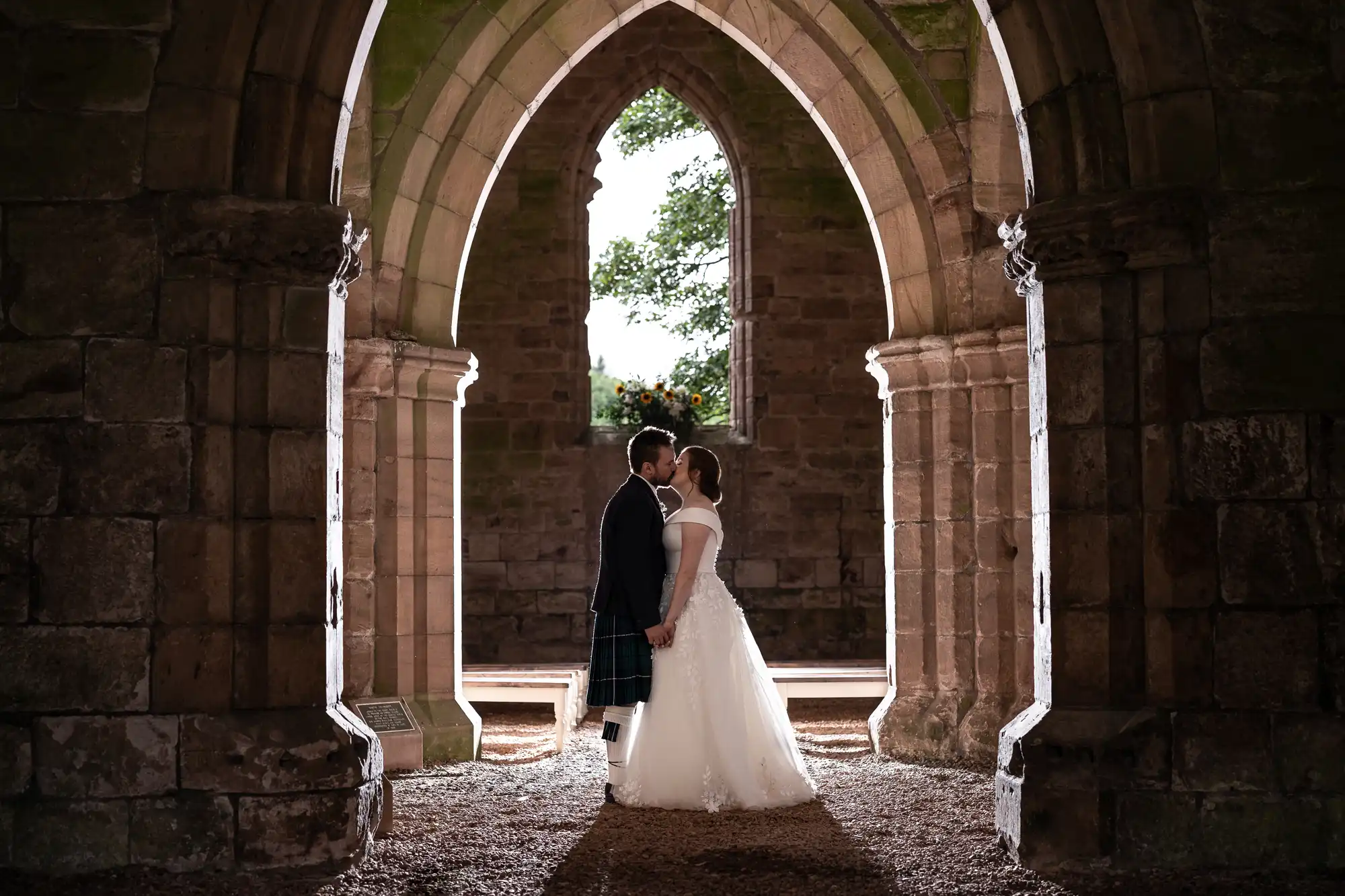 A couple in wedding attire holds hands and kisses inside a stone archway of a historic building, bathed in soft light from an opening in the background.