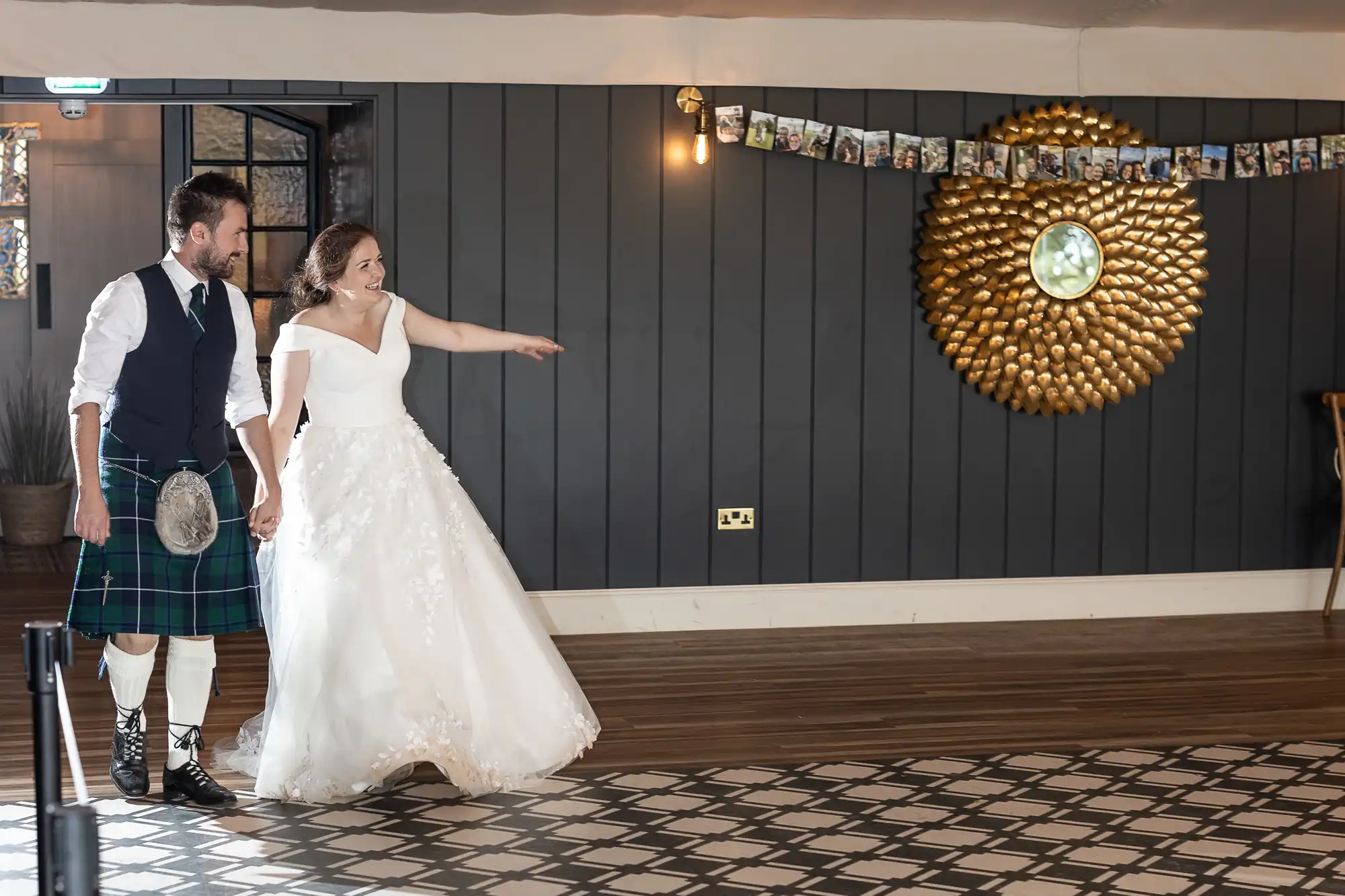 A bride and groom, with the groom in a kilt, hold hands and look in the same direction inside a room with dark-paneled walls and a decorative wall piece. A photo banner is hung above them.