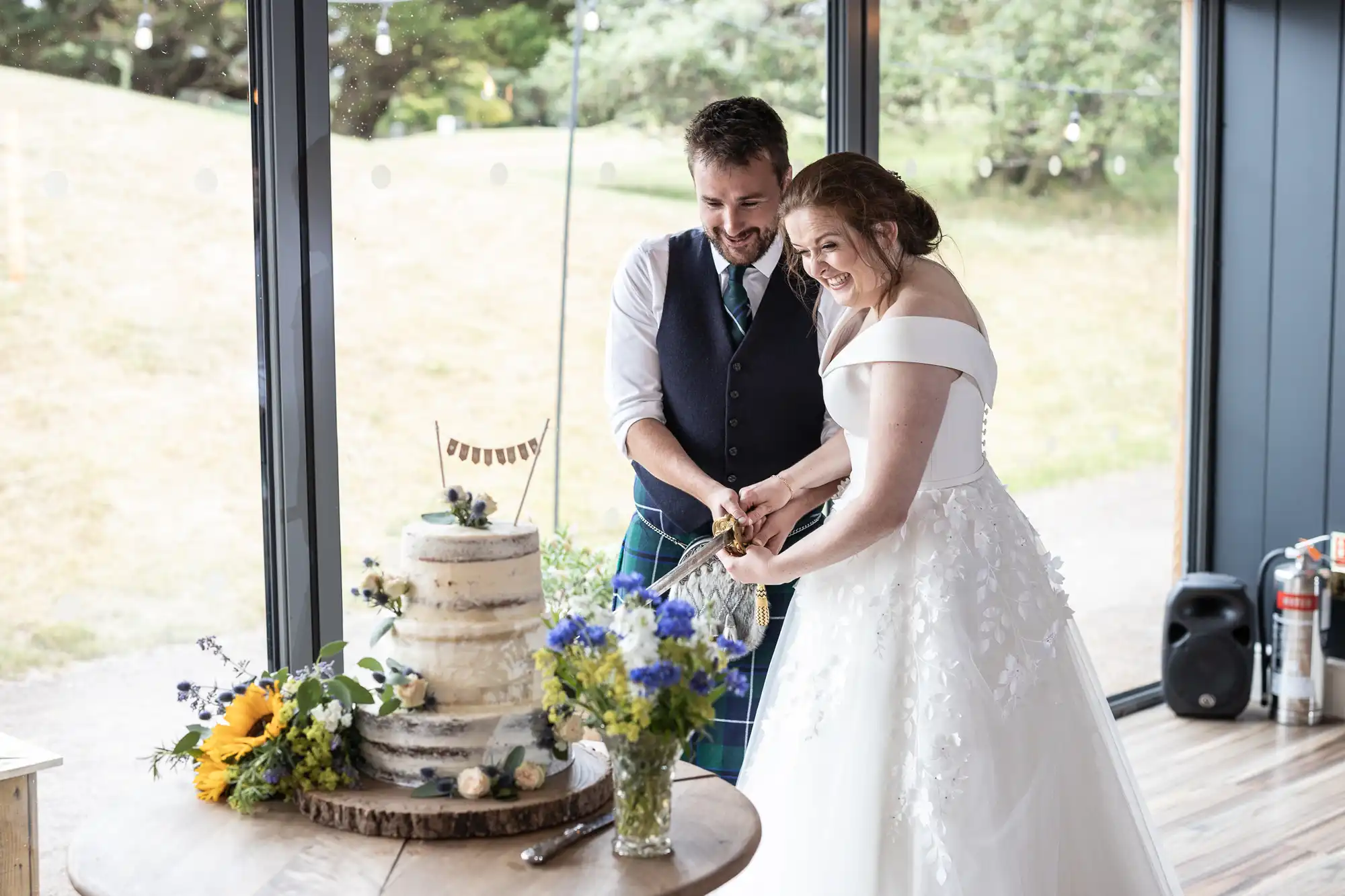 A bride and groom, dressed in formal wedding attire, smile as they cut their wedding cake together in a room with large windows and a view of greenery outside.