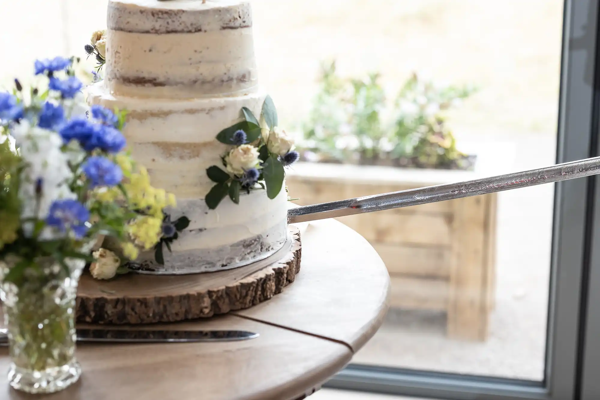 A two-tier rustic cake being cut with a large sword; a decorated vase of flowers is placed next to it on a wooden surface.