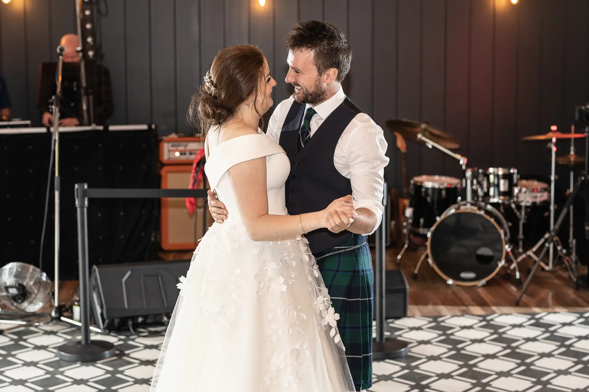 A couple dressed in wedding attire dances together in a venue with musical instruments in the background.