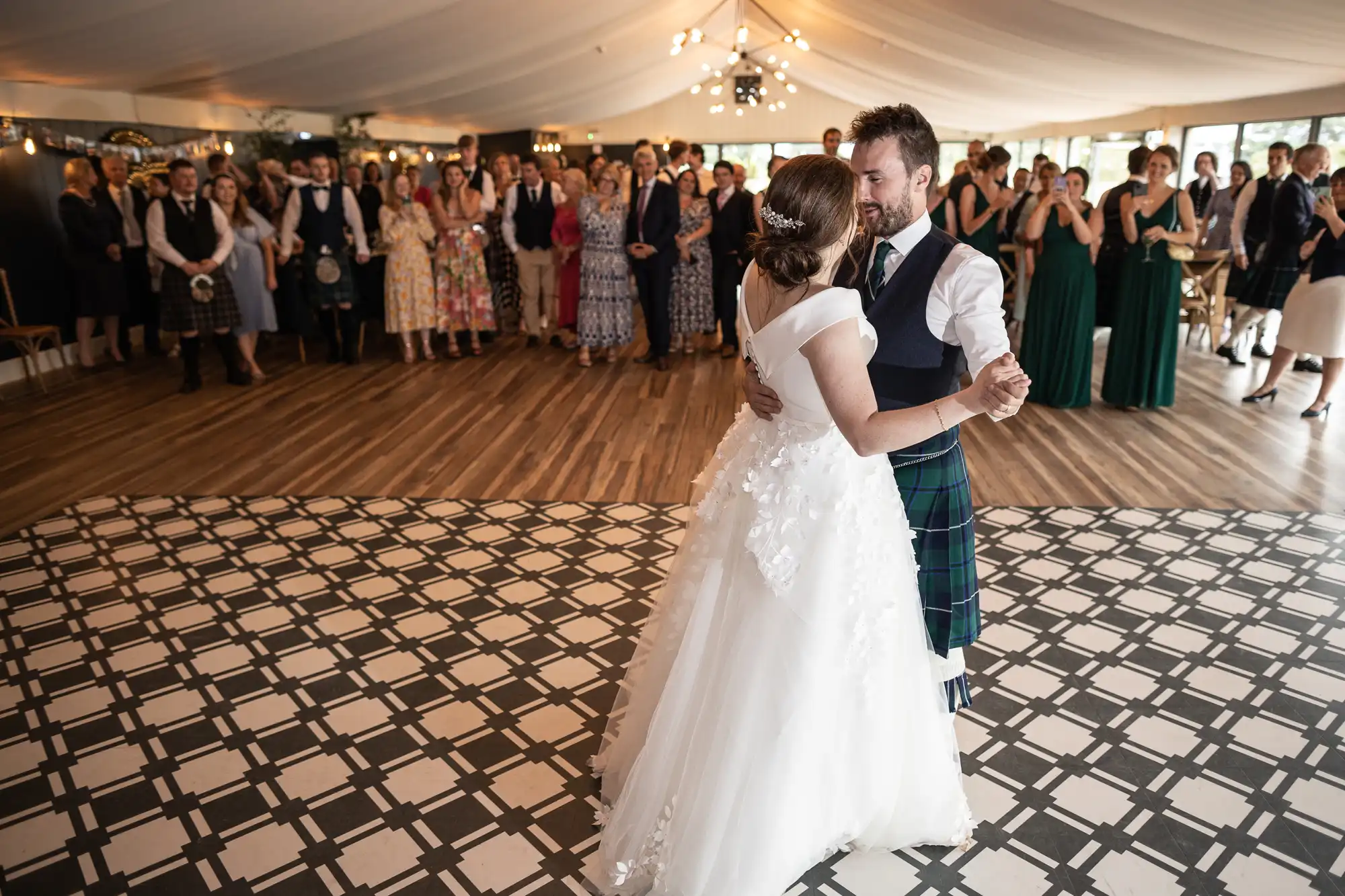 A couple is dancing together on a patterned floor while formally dressed guests look on in a large, decorated indoor space.