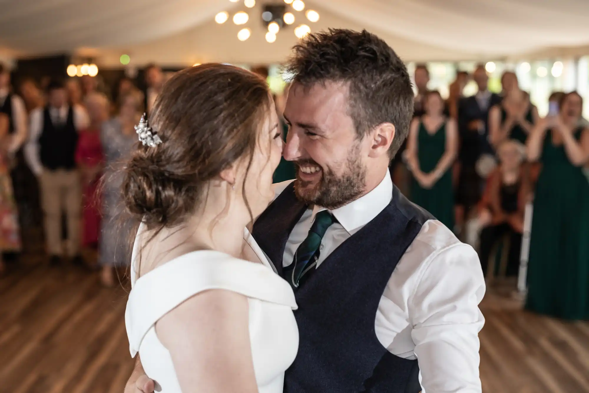 A couple dressed in formal attire shares a joyful moment on the dance floor, surrounded by smiling onlookers in a well-lit, decorated venue.