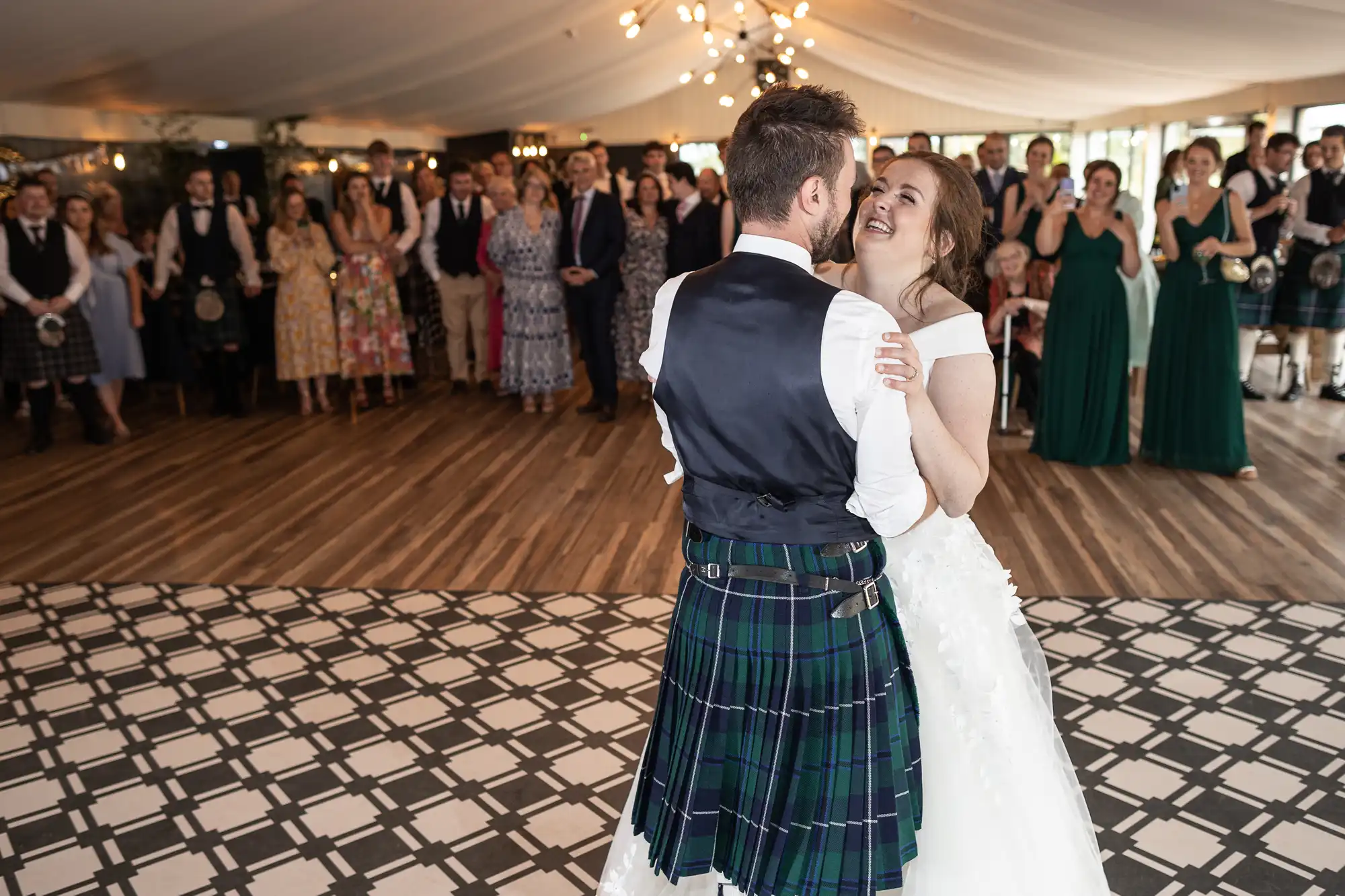 A bride and groom share their first dance at a wedding reception, with onlookers standing and watching in the background. The groom wears a traditional kilt.