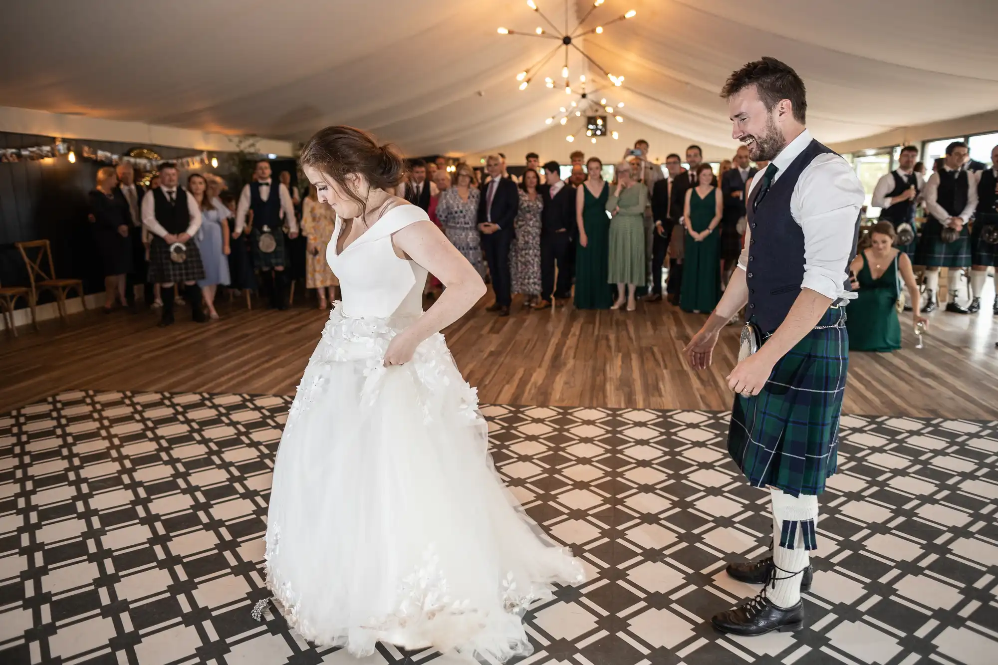 A couple dressed in wedding attire dances on a patterned floor in a tented venue, with guests watching in the background.