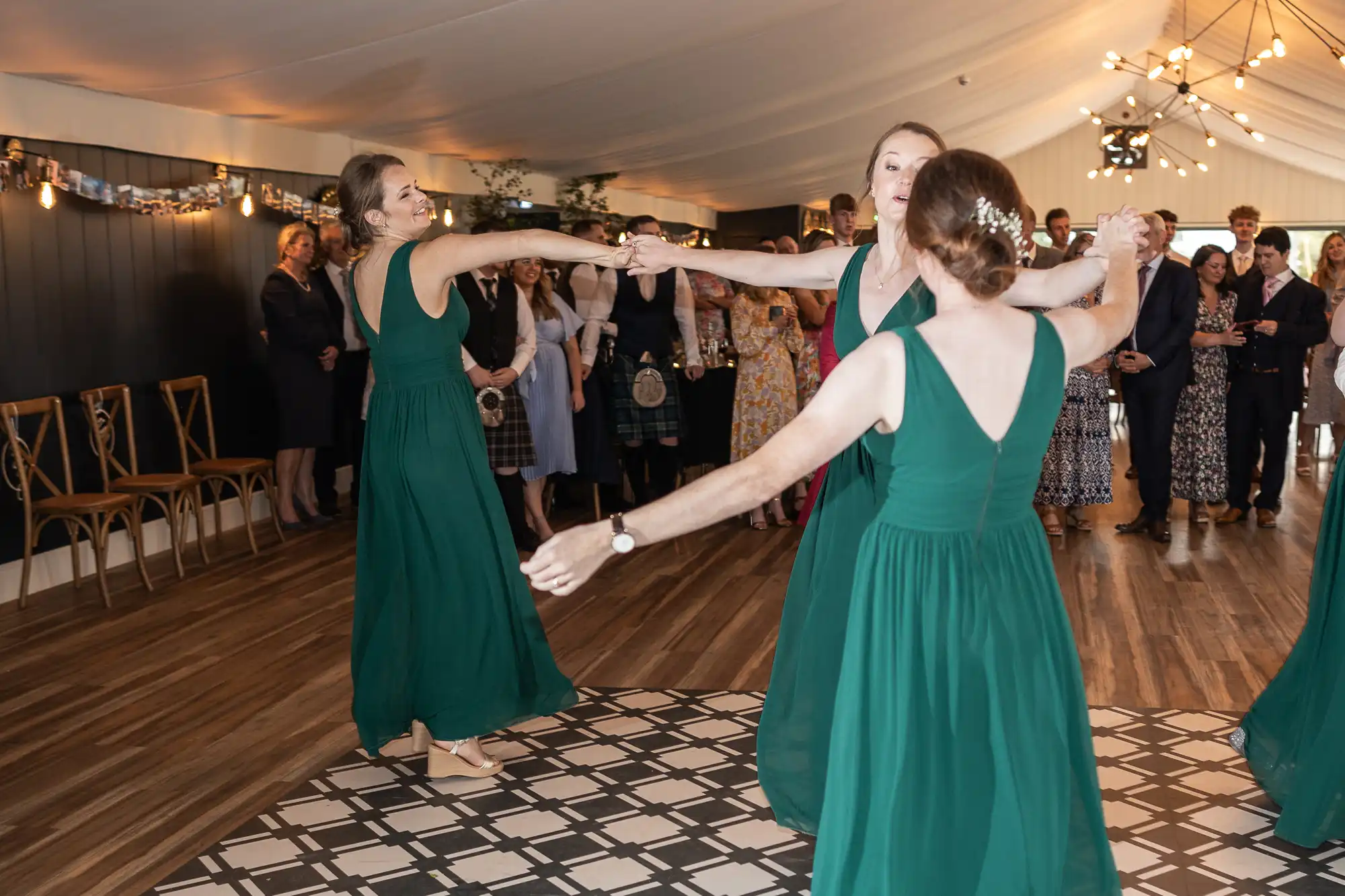 Three women in green dresses dance in a circle at an indoor wedding reception, while guests stand in the background watching.