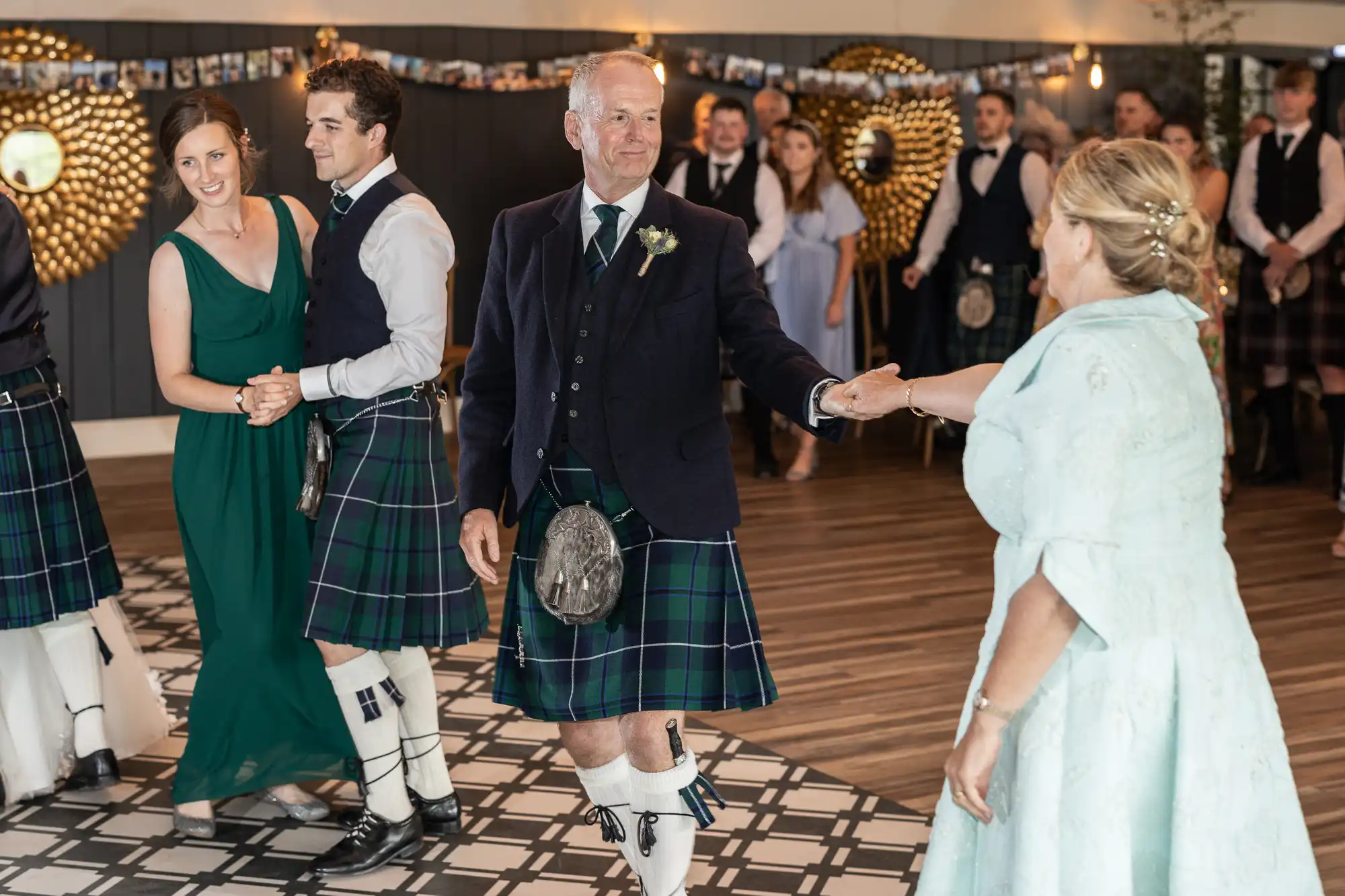 Guests in Scottish attire dance at a wedding reception, with men wearing kilts and women in formal dresses. Decorations include string lights and a photo collage in the background.