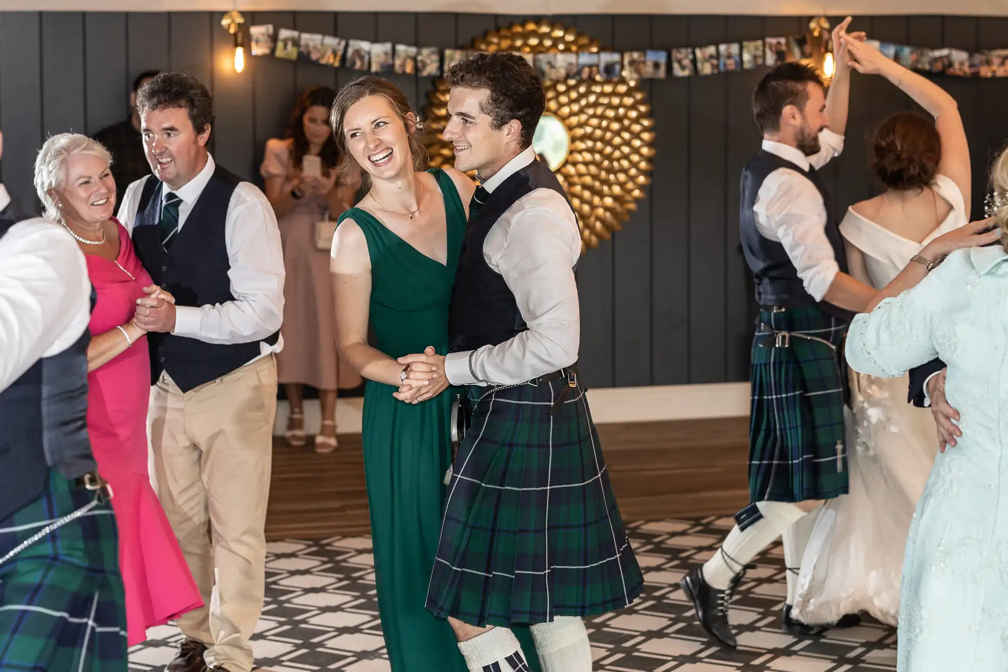 Guests dancing in pairs at an event, with some men wearing kilts and women dressed in dresses. A decorative mirror and photo garland are visible in the background.