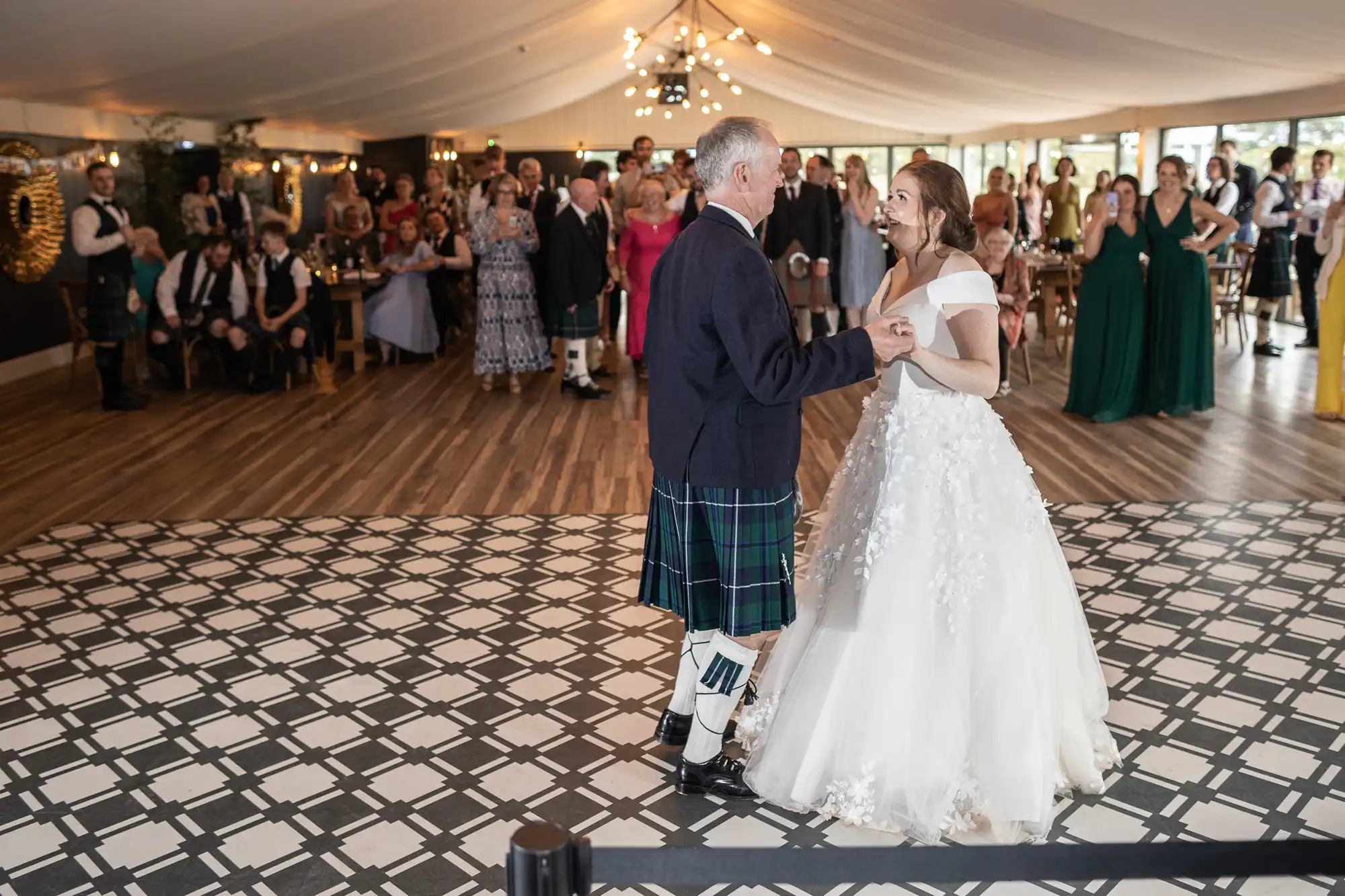 A bride in a white dress dances with an elderly man in a kilt on a patterned floor, with a group of seated guests watching in a tented venue.